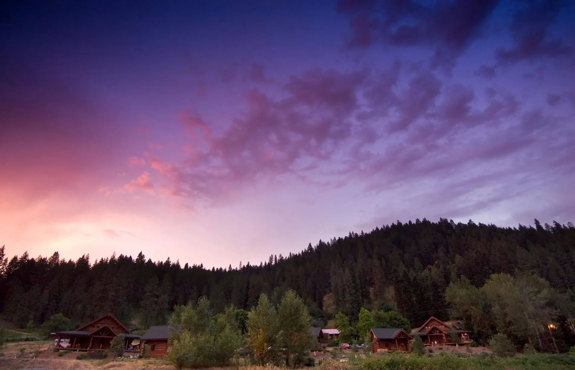 Log cabins nestled in a forest under a vibrant purple and orange sunset sky.