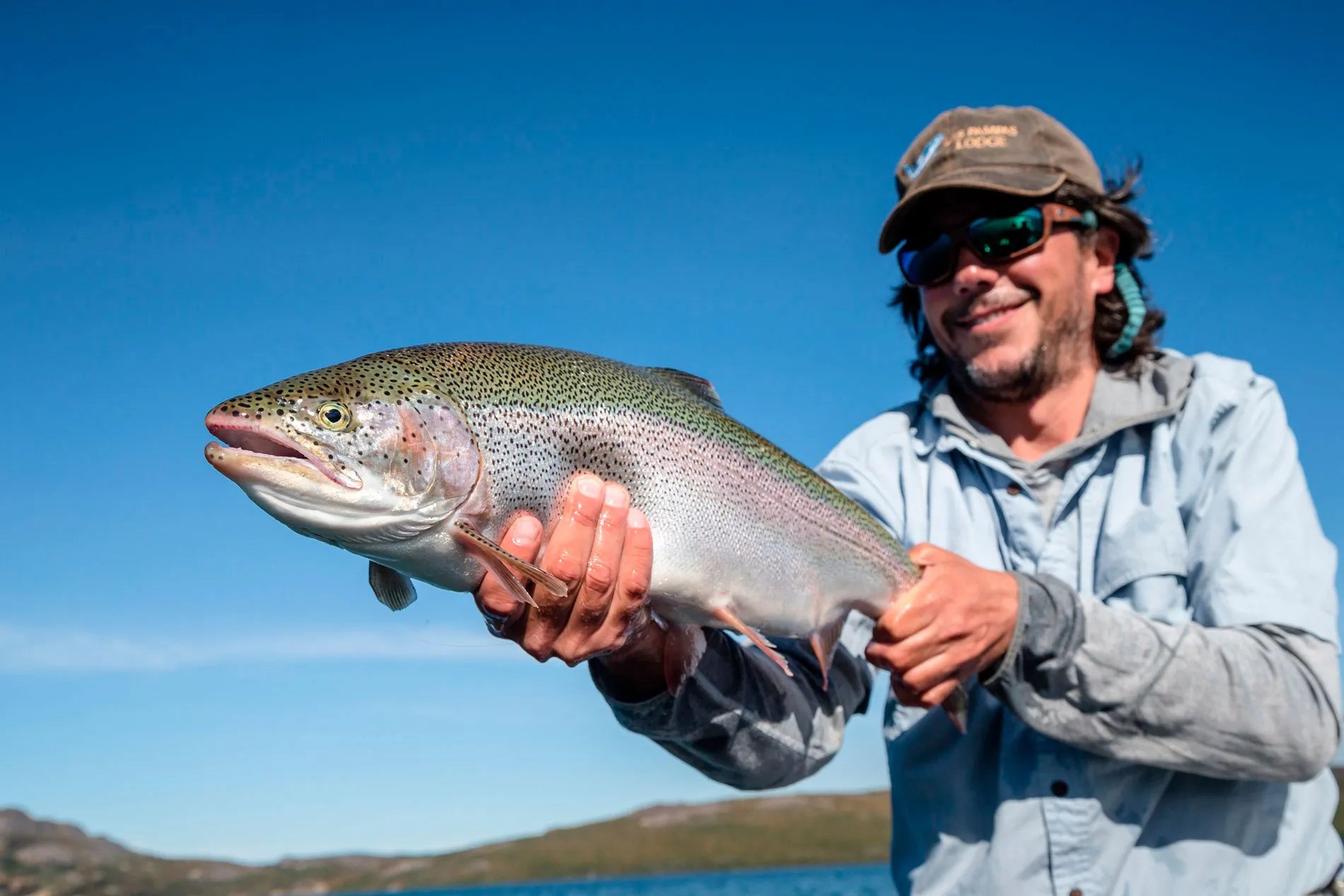 Smiling angler holding a large, iridescent rainbow trout against a bright blue sky.