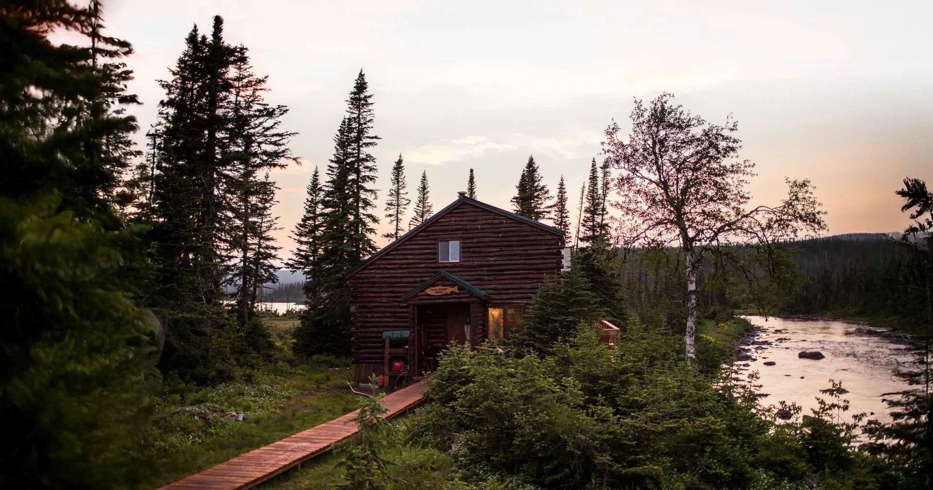 Rustic log cabin lodge nestled among trees by a river at sunset.