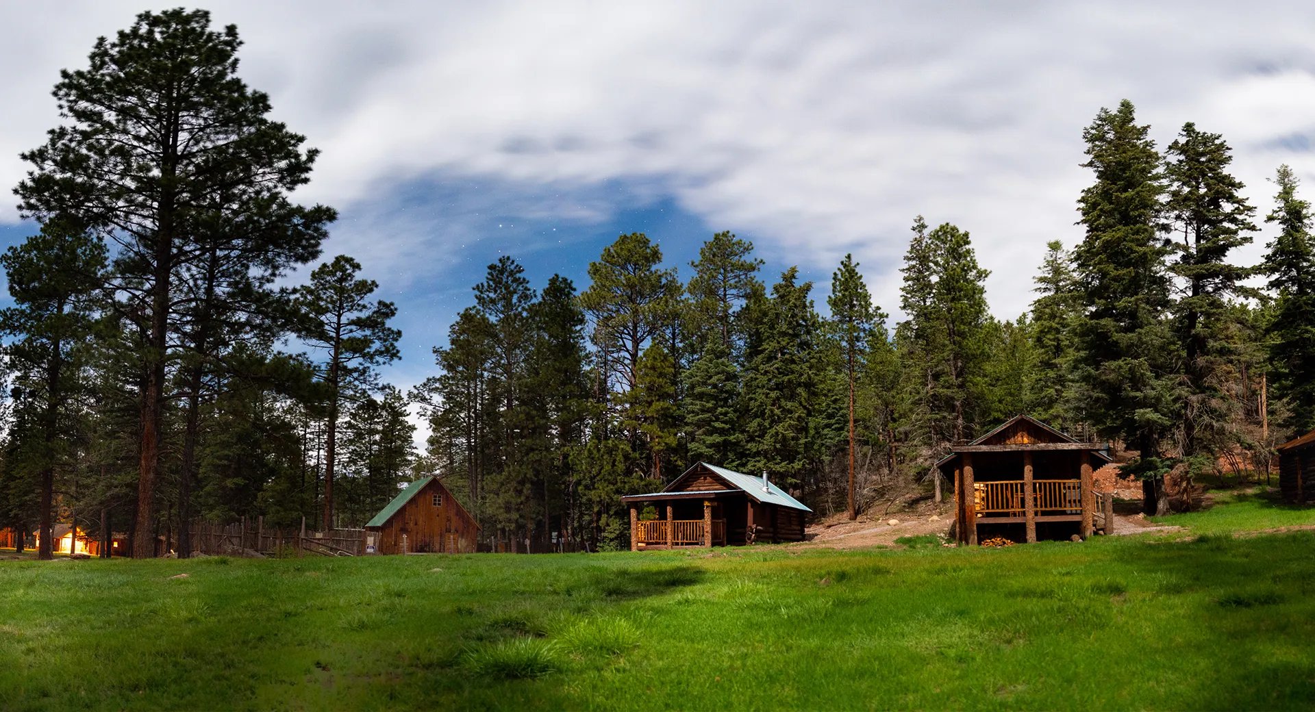 Grassy meadow with rustic log cabins and tall pine trees under a partly cloudy blue sky.