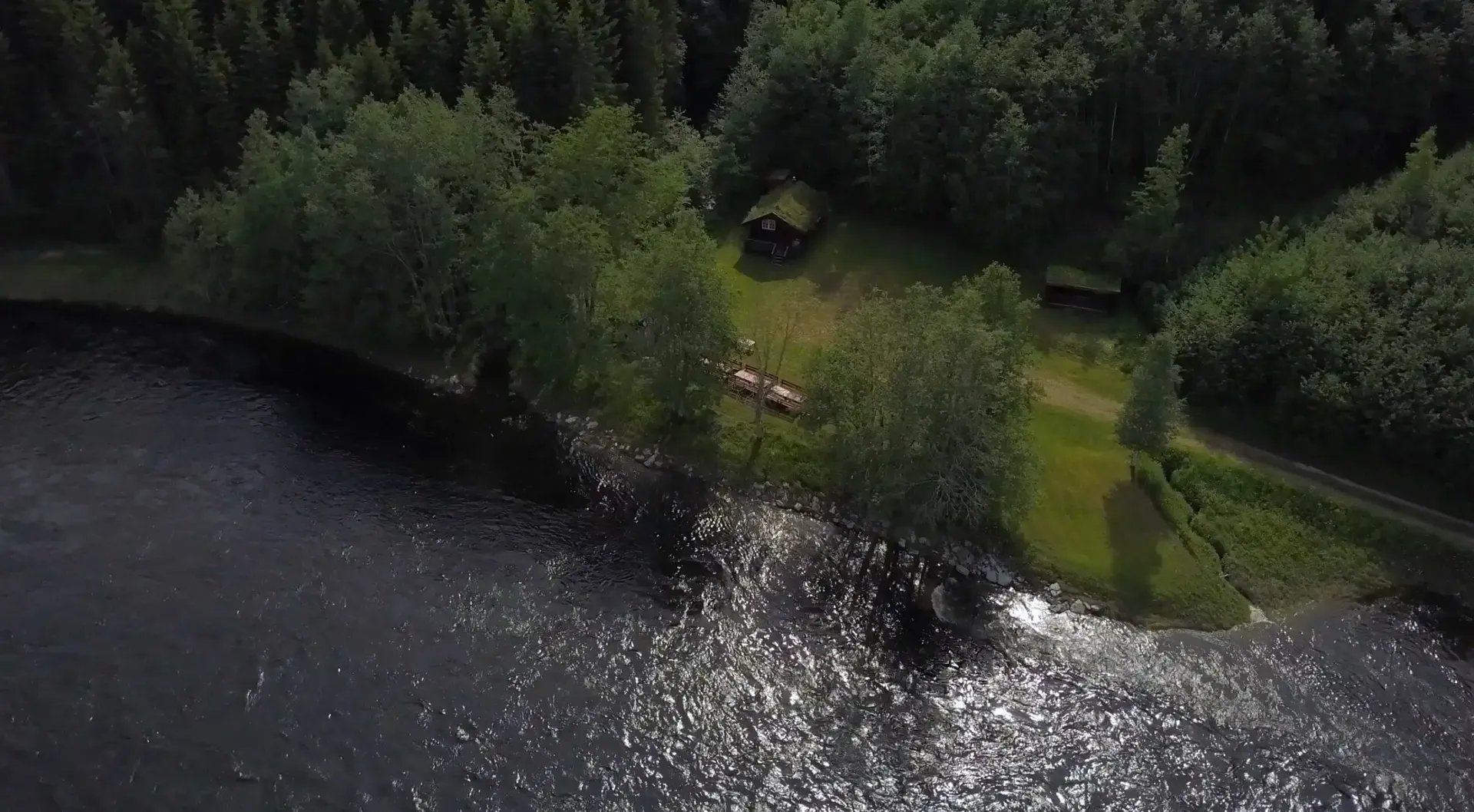 Aerial view of a forested riverbank with small cabins and a path