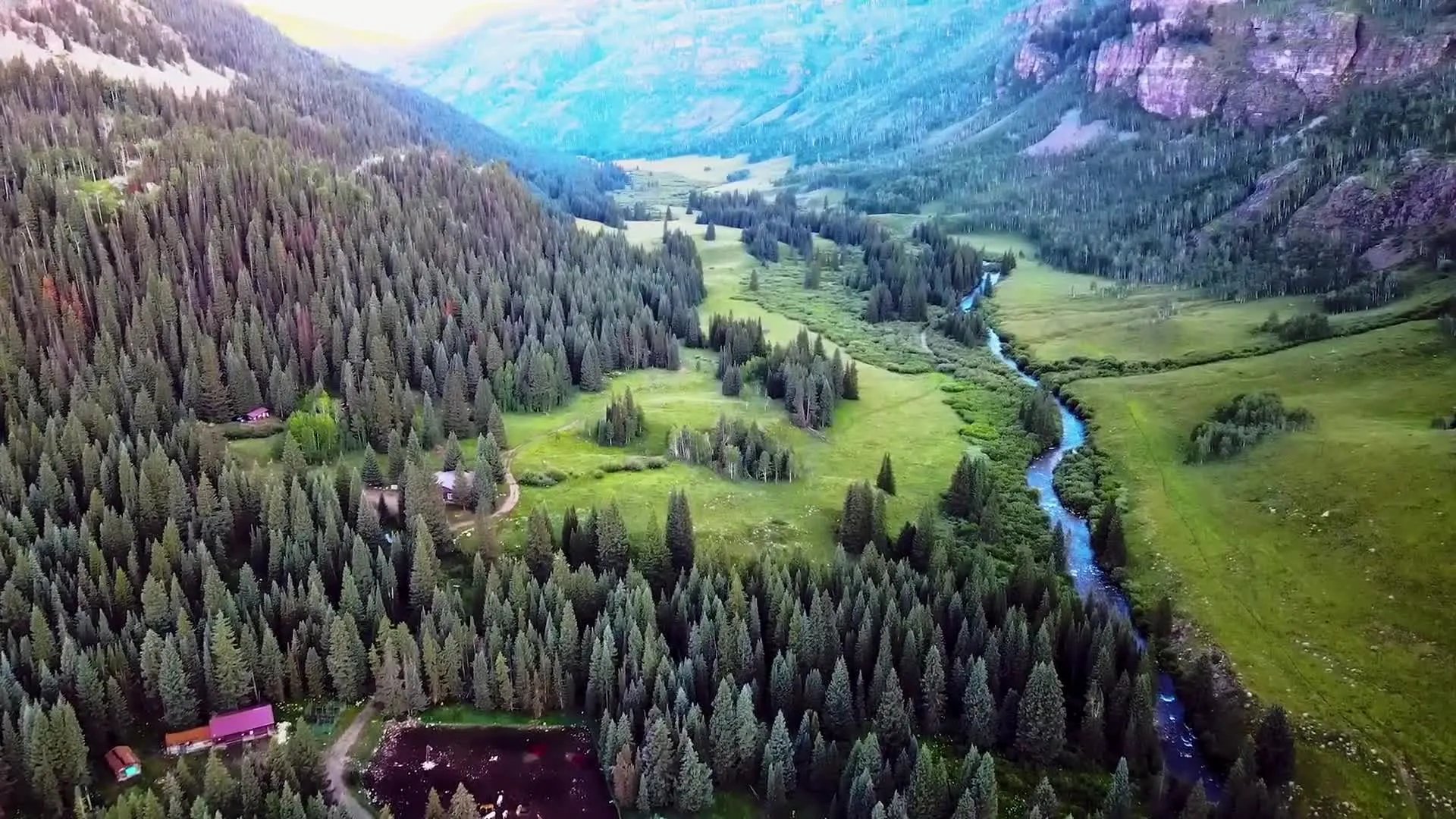 Aerial view of a lush green mountain valley with a winding river and scattered lodge buildings.