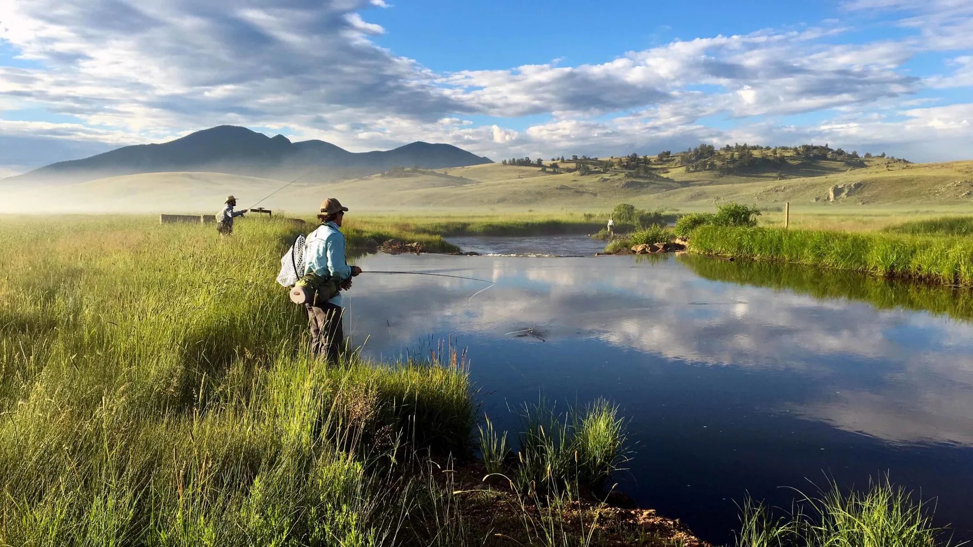 Angler casting a fly rod in a misty river at sunrise with mountains in the background