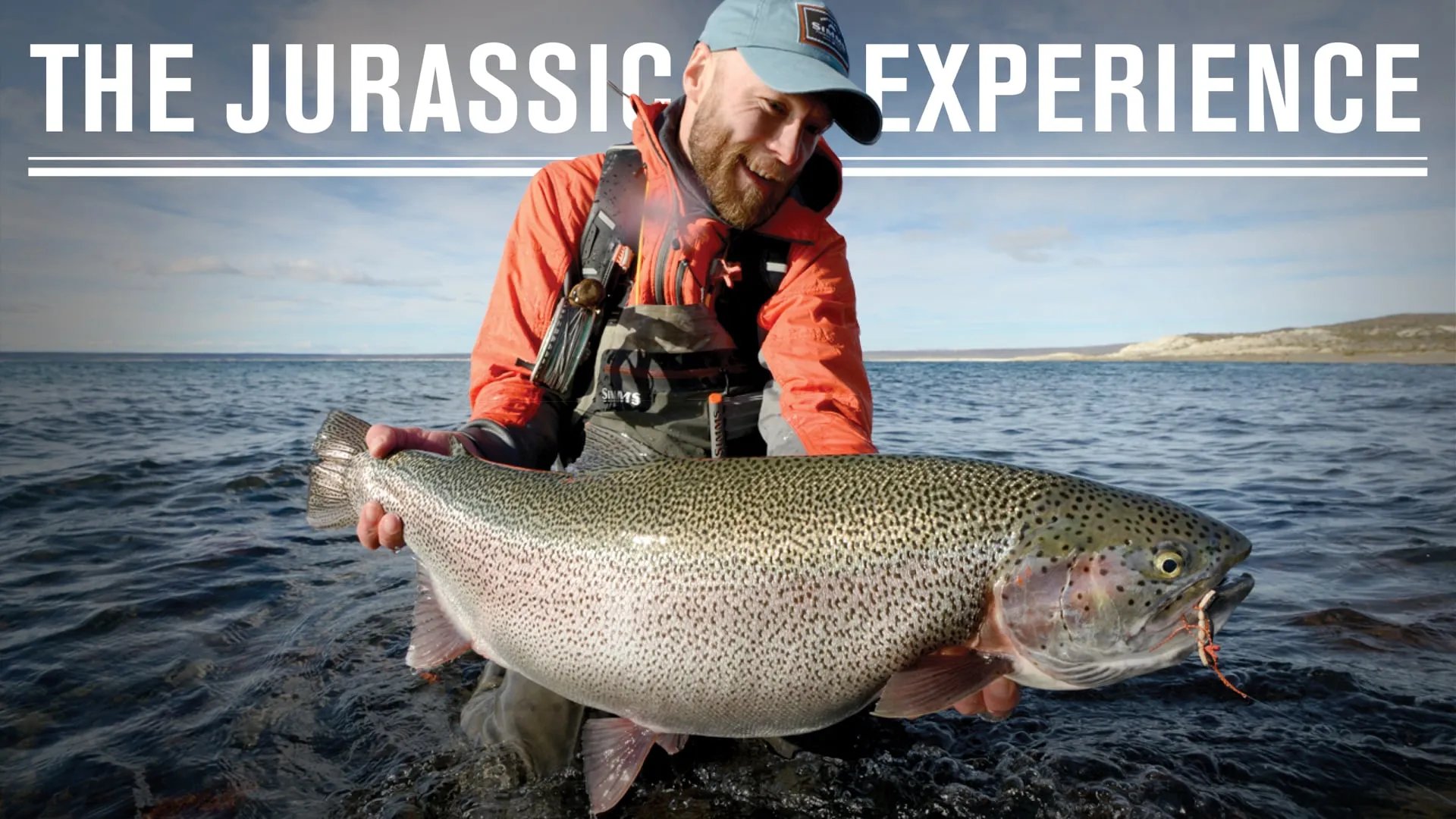 Angler in waders holding a large rainbow trout in shallow water under a cloudy sky