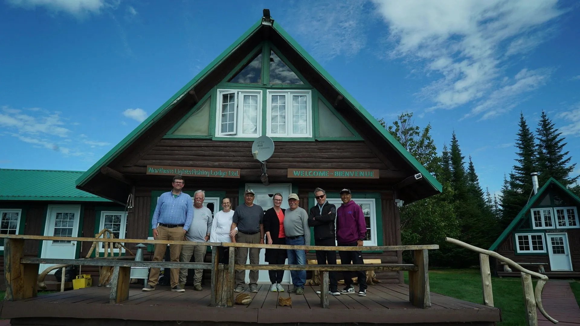 Group of people standing on the wooden porch of a large A-frame lodge building under a blue sky