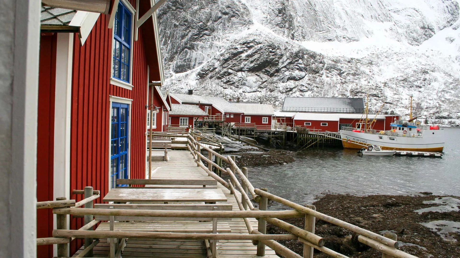 Snowy red rorbu cabins on a wooden boardwalk next to a frozen fjord with boats and snow-capped mountains
