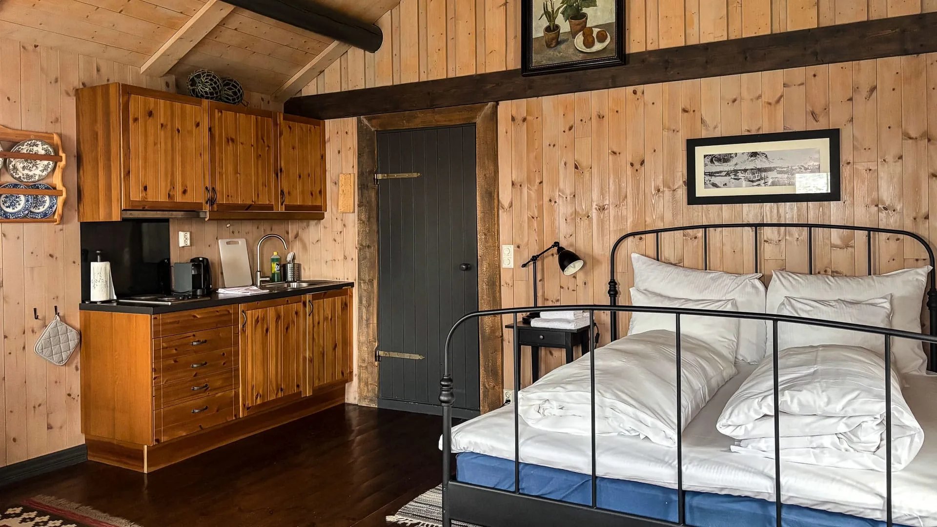 Bedroom with warm wood paneling, a black metal-frame double bed, and a compact kitchenette