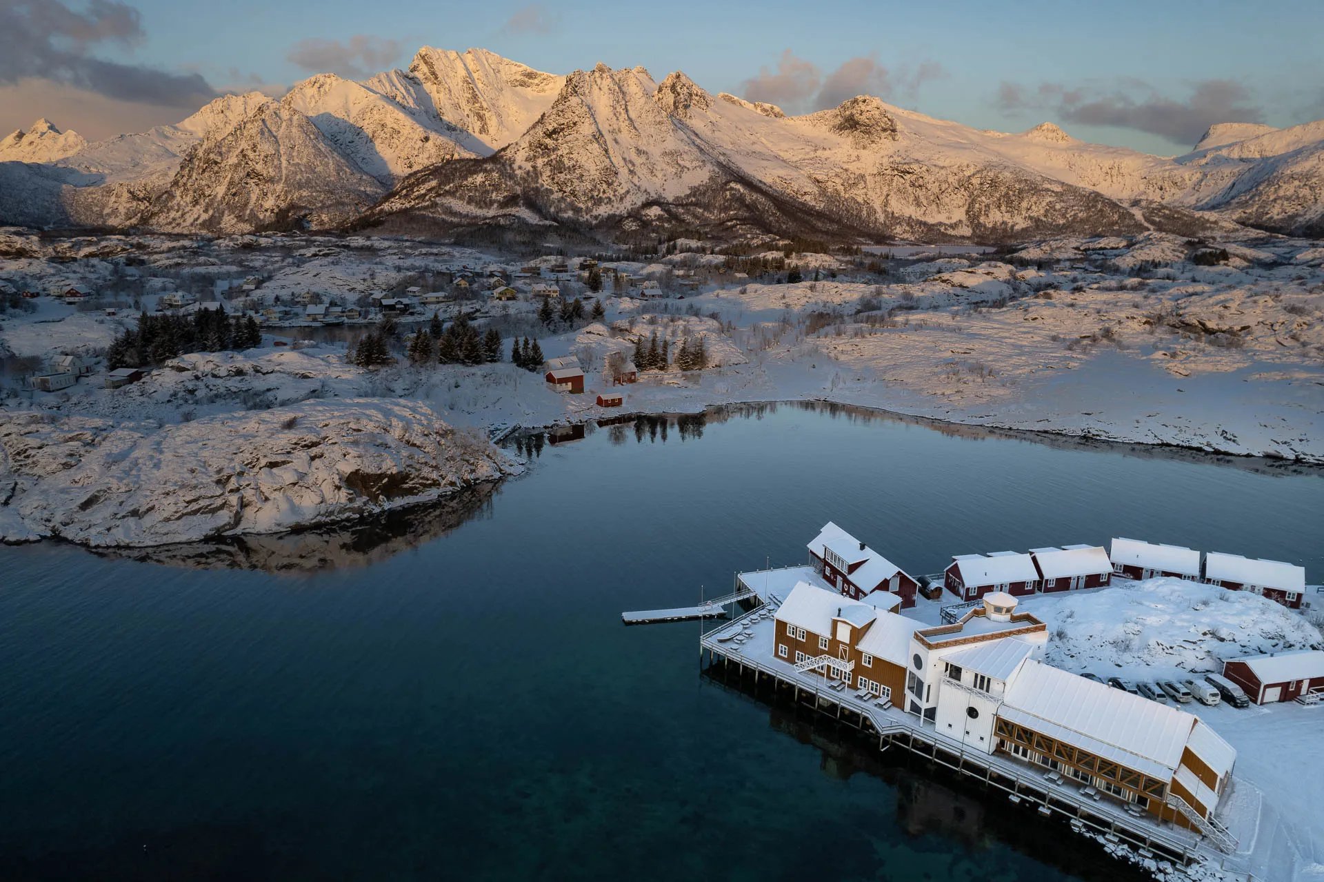 Aerial view of Lofoten Ski Lodge complex with red cabins and main building on a snowy waterfront, surrounded by mountains