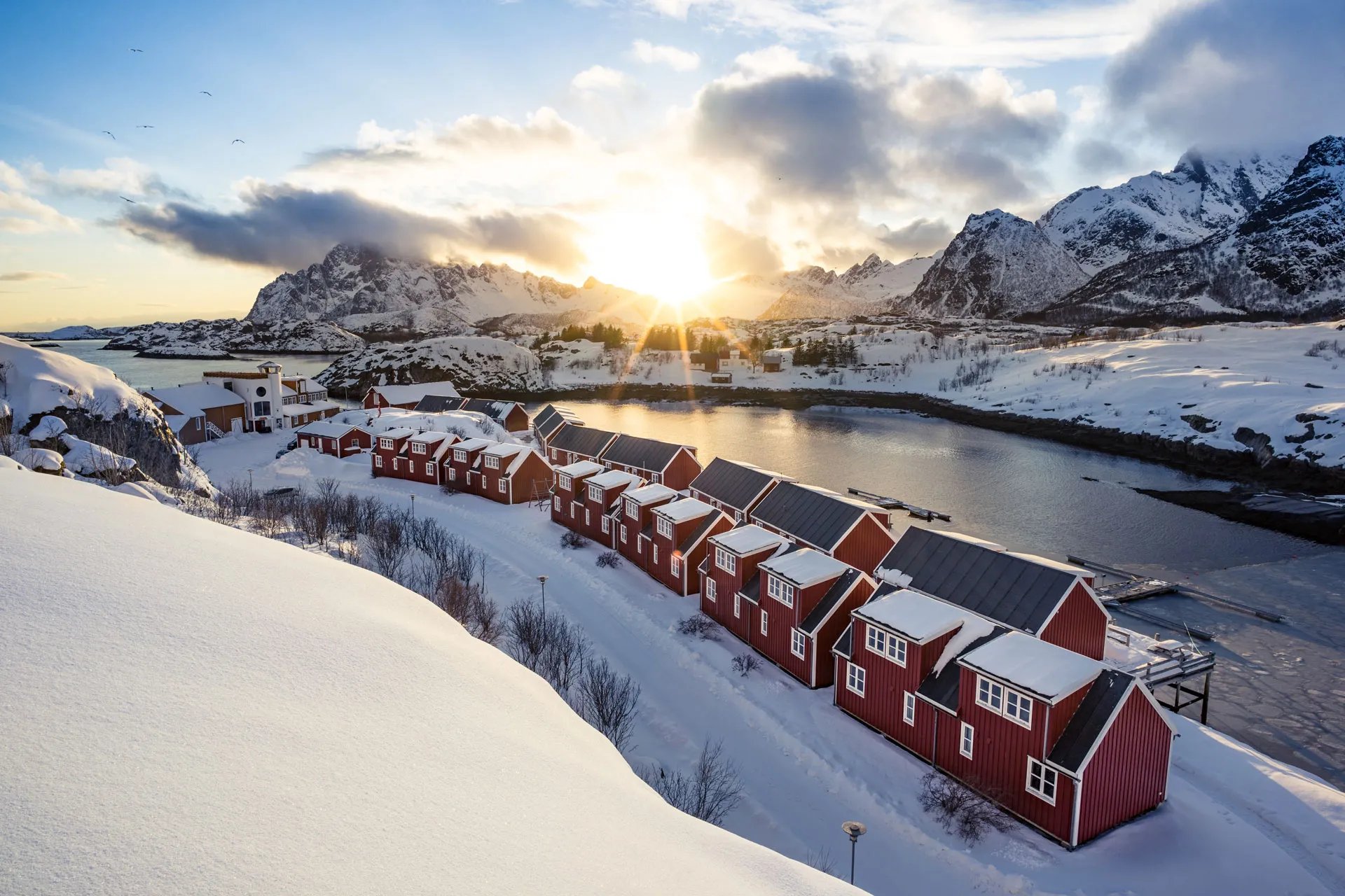 Row of traditional red cabins along a snowy shoreline at sunset, with mountains in the background