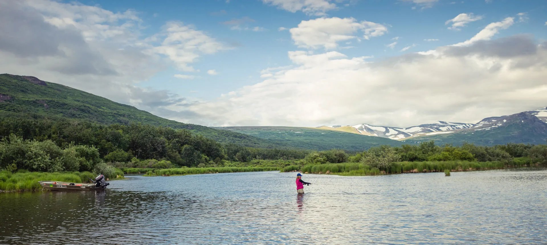 Two anglers wading in a wide river, surrounded by green hills and distant snow-capped mountains.