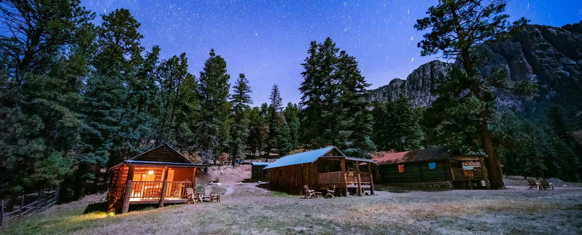 Rustic log cabins nestled among tall pine trees under a starry night sky.