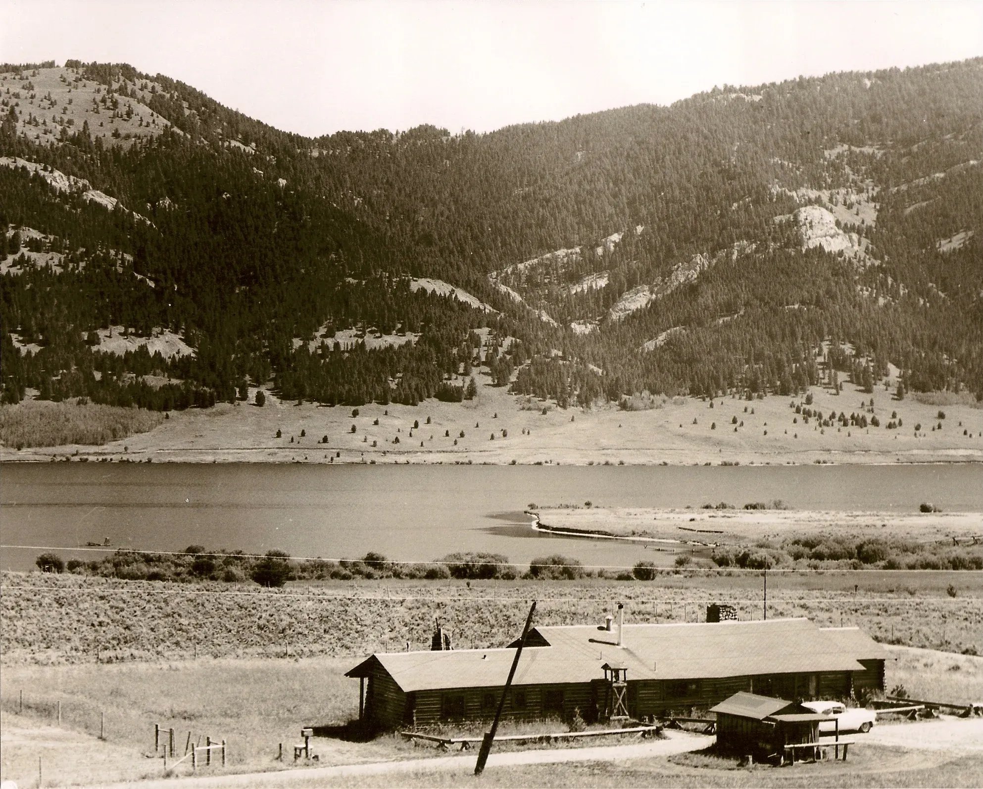 Historic sepia-toned view of the main lodge building by a lake with mountains in the background.