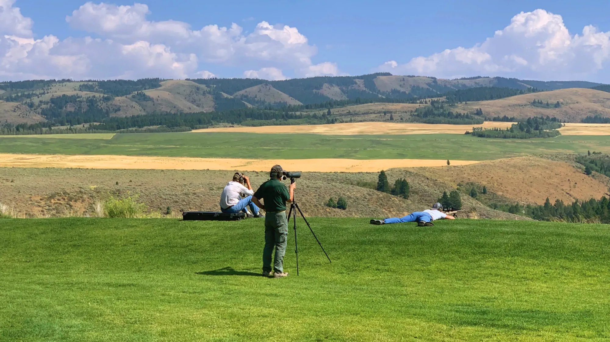 People on a grassy hill with a spotting scope, overlooking a vast, green and golden valley