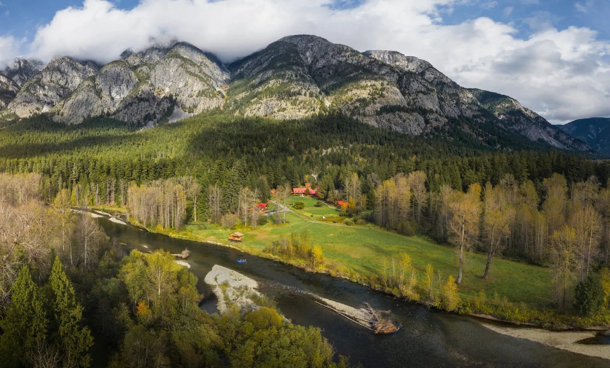 Aerial view of Tweedsmuir Park Lodge in autumn, nestled beside a winding river with rafts on the water, surrounded by golden deciduous trees, evergreen forest, and dramatic snow-dusted mountain peaks under low clouds.