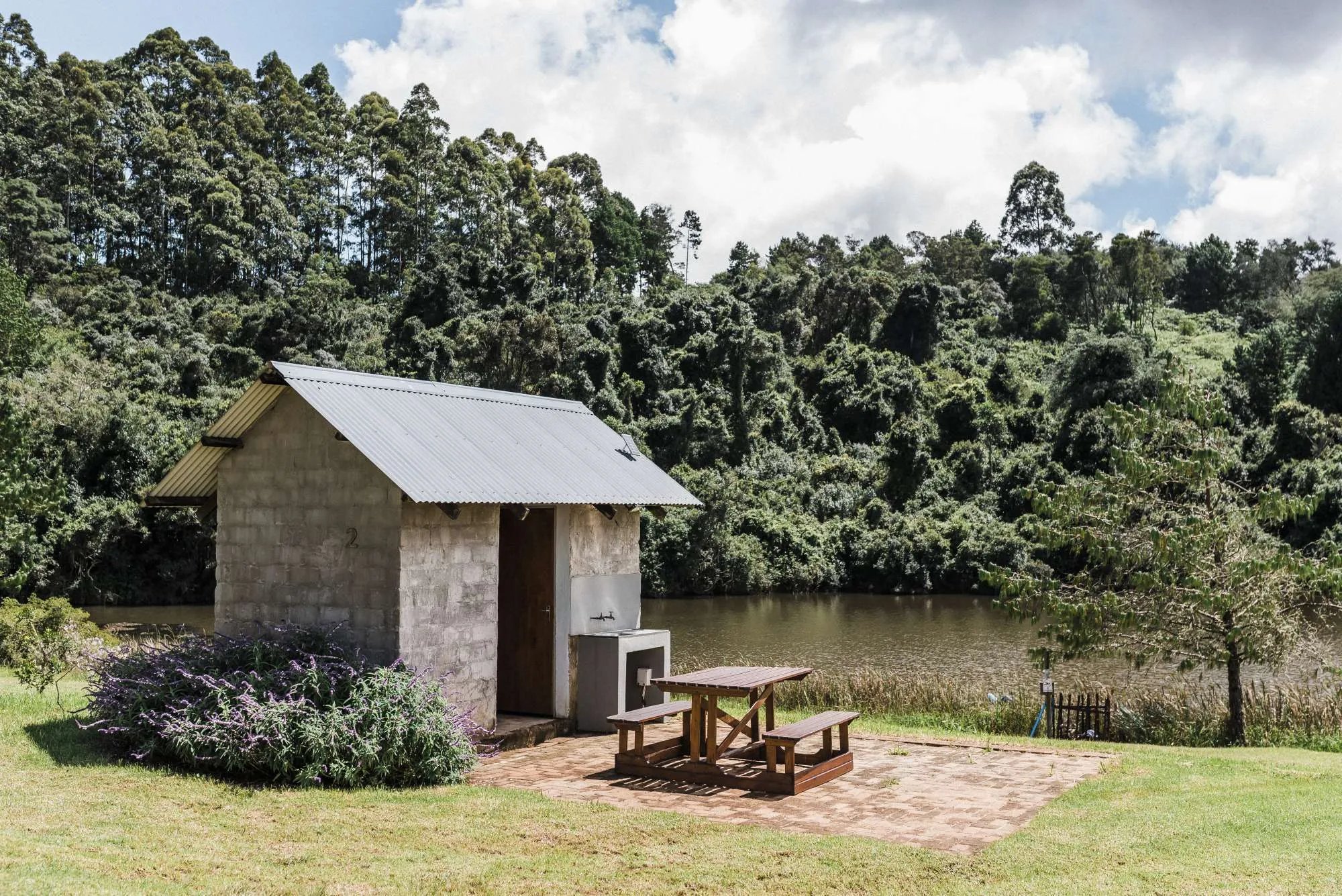 Small stone building with a metal roof and a wooden picnic table by a lake.