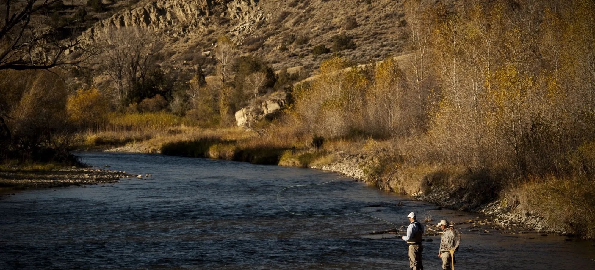 Two anglers standing in a river with a rocky cliff and autumn trees in the background.