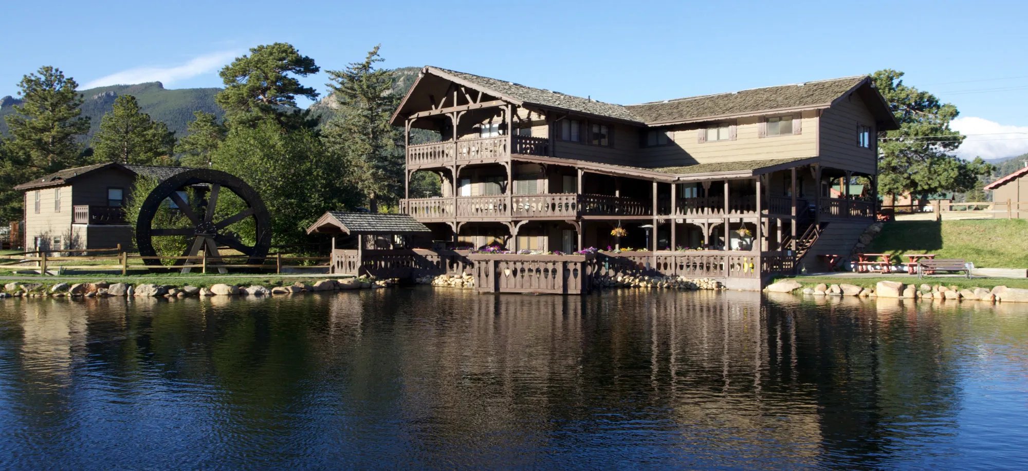 Large multi-story lodge building with a water wheel on a calm pond, reflecting the structure.