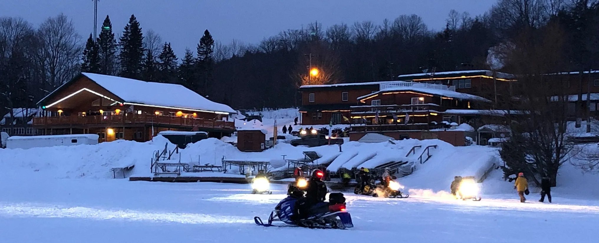 AJ's Lodge & Oven buildings illuminated at night in a snowy winter landscape with snowmobiles