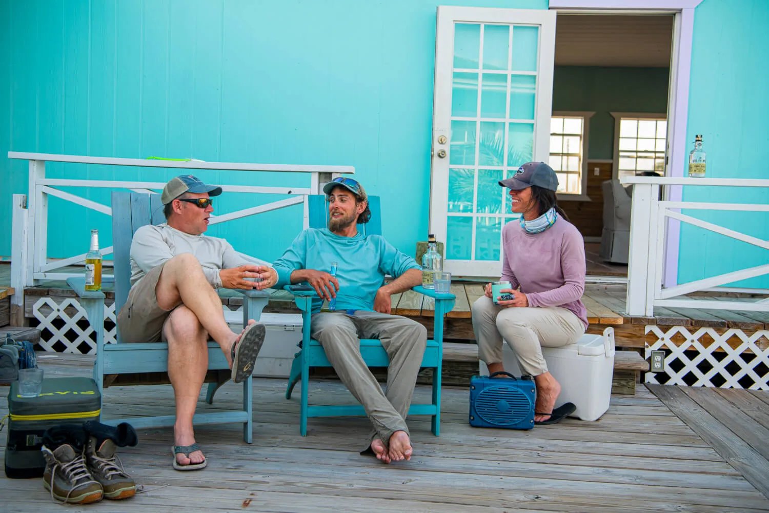 Three people relaxing in Adirondack chairs on a wooden porch of a turquoise lodge building