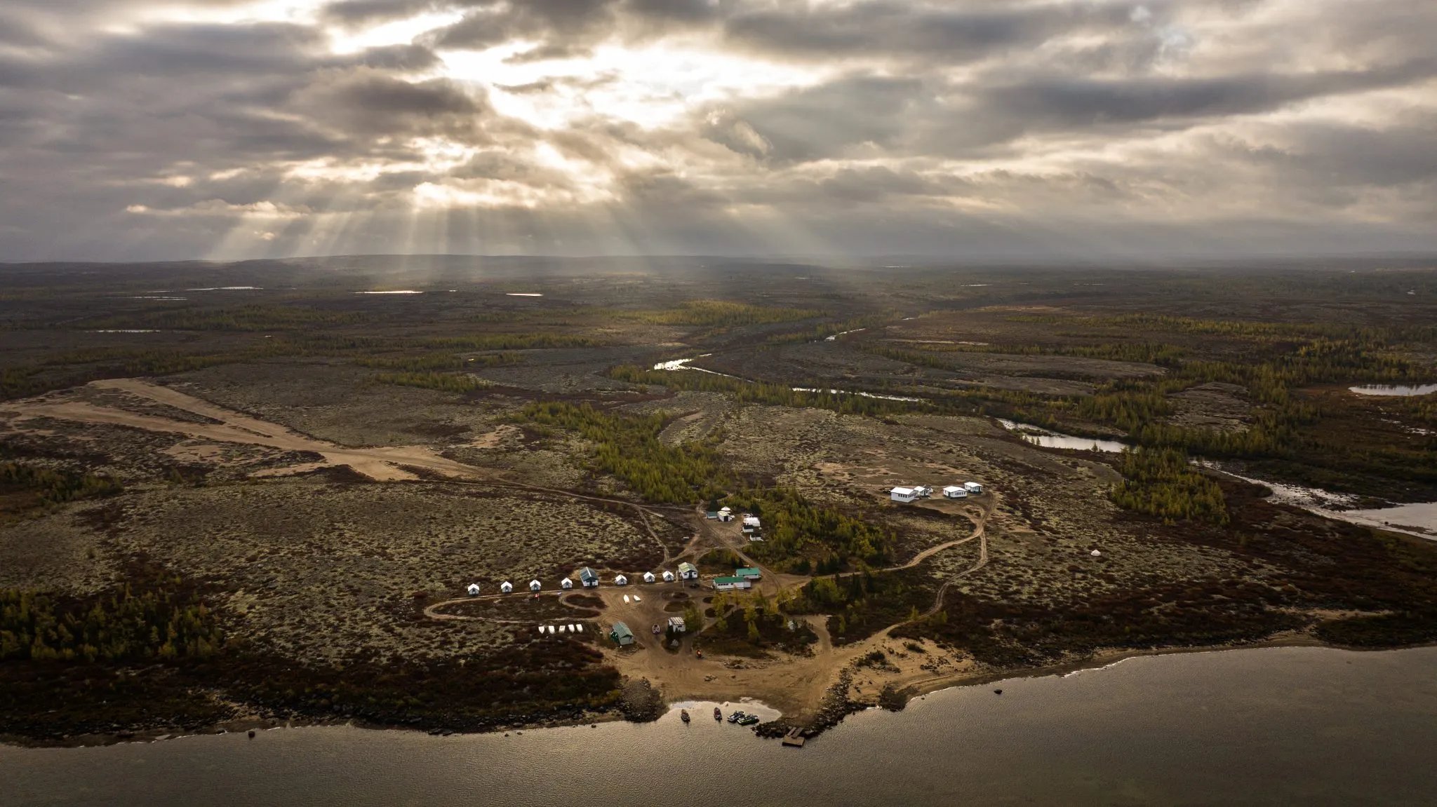 Aerial view of Leaf River Lodge camp by a river under dramatic, sun-dappled clouds