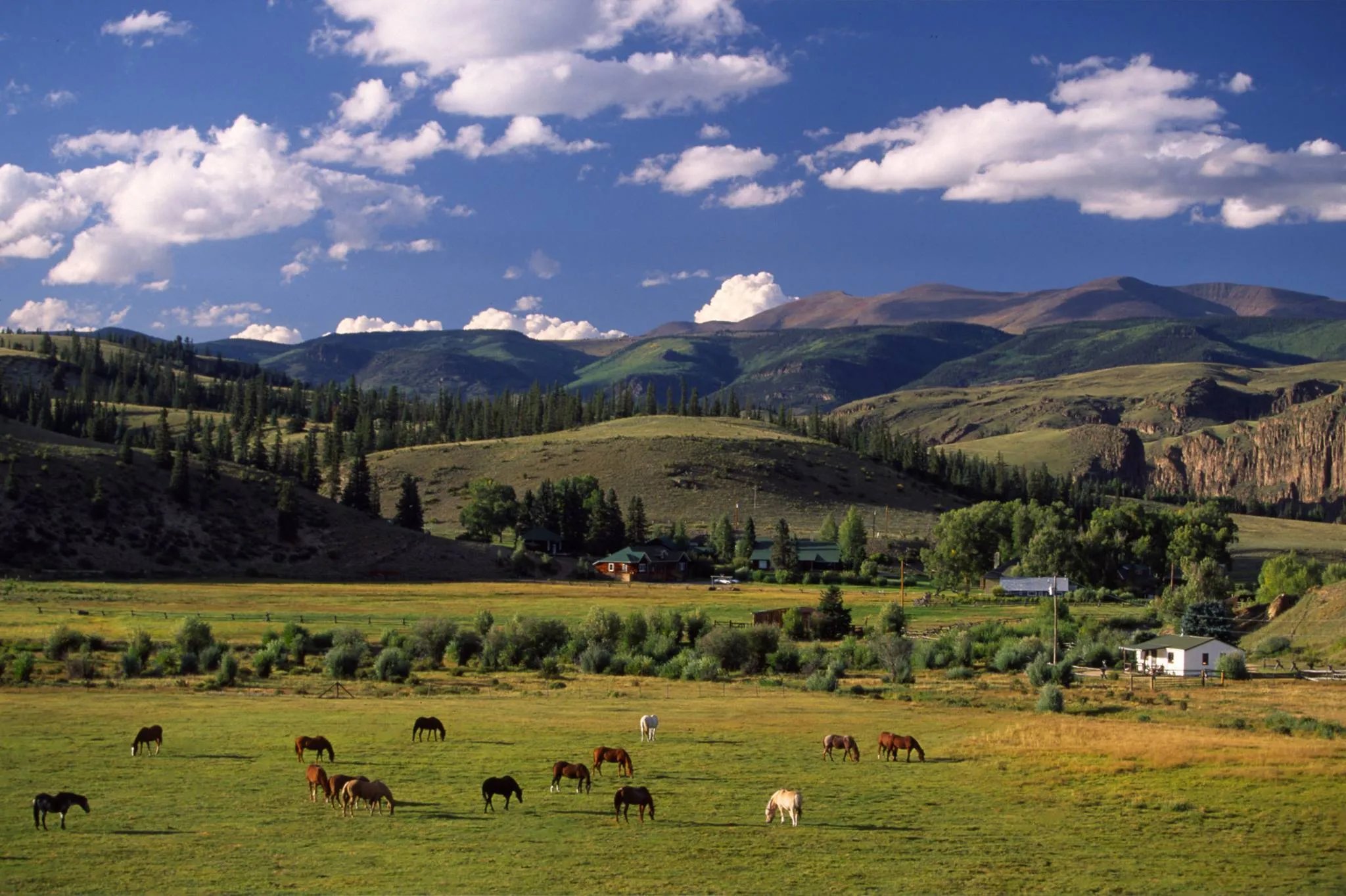 Panoramic view of a green mountain valley with horses grazing and ranch buildings in the distance.