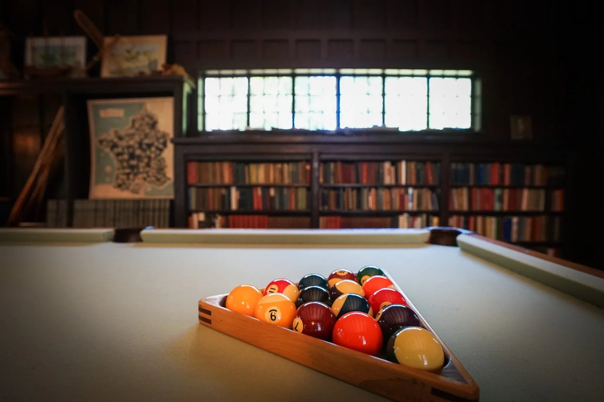 Pool table with billiard balls arranged in rack in a dimly lit room with bookshelves and a window in the background.
