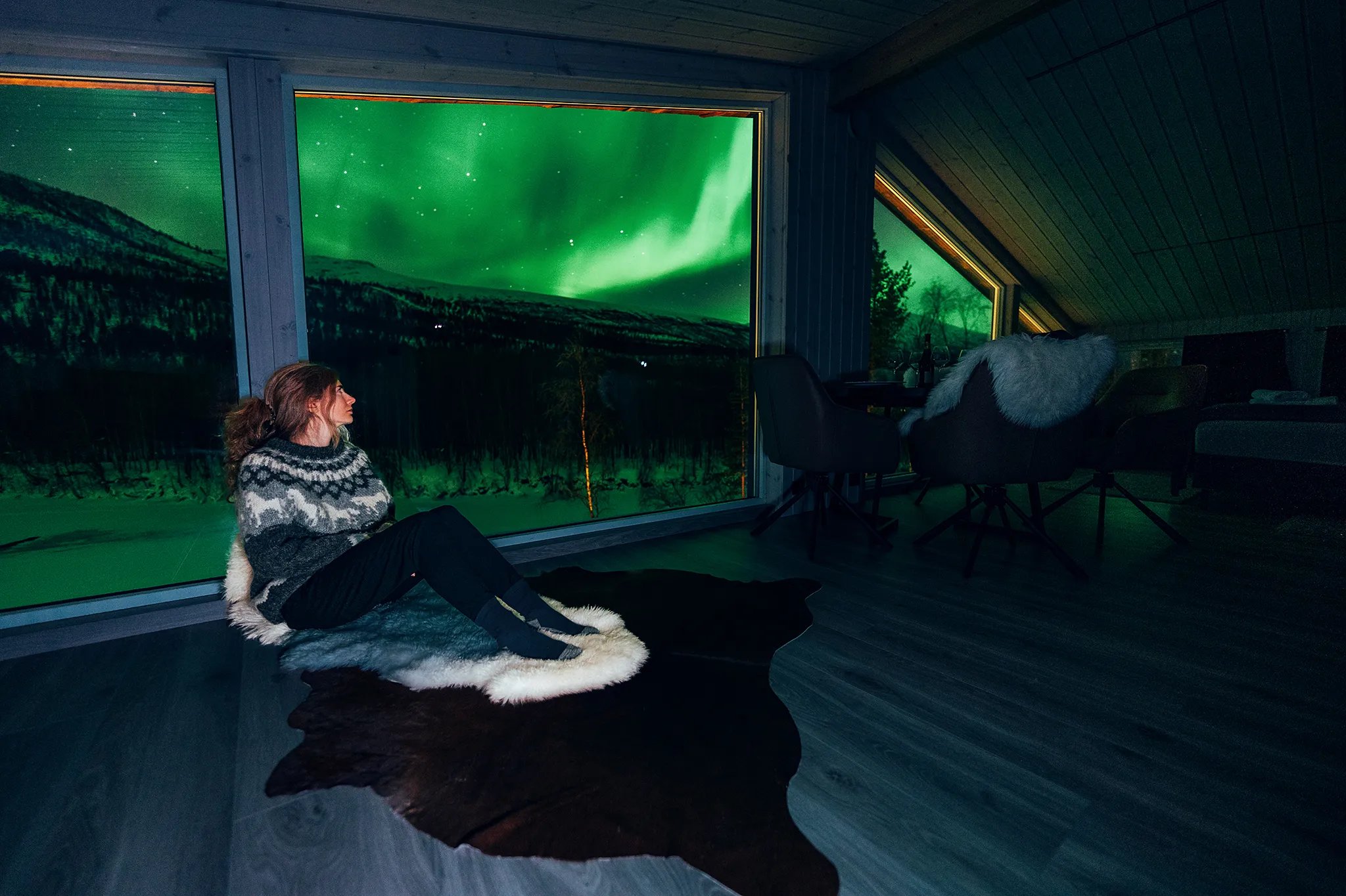 Woman in a cozy sweater watching green Northern Lights from a large window in a lodge room.