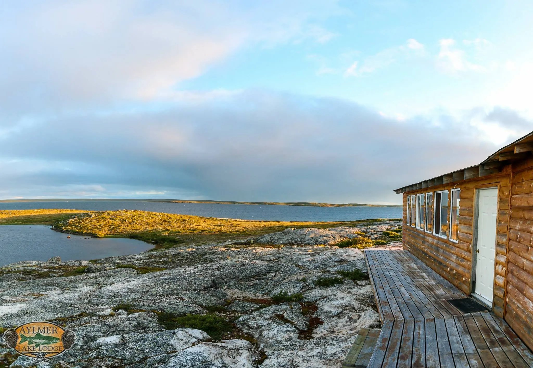 Rustic wooden cabin with a deck overlooking a lake and tundra landscape at dusk