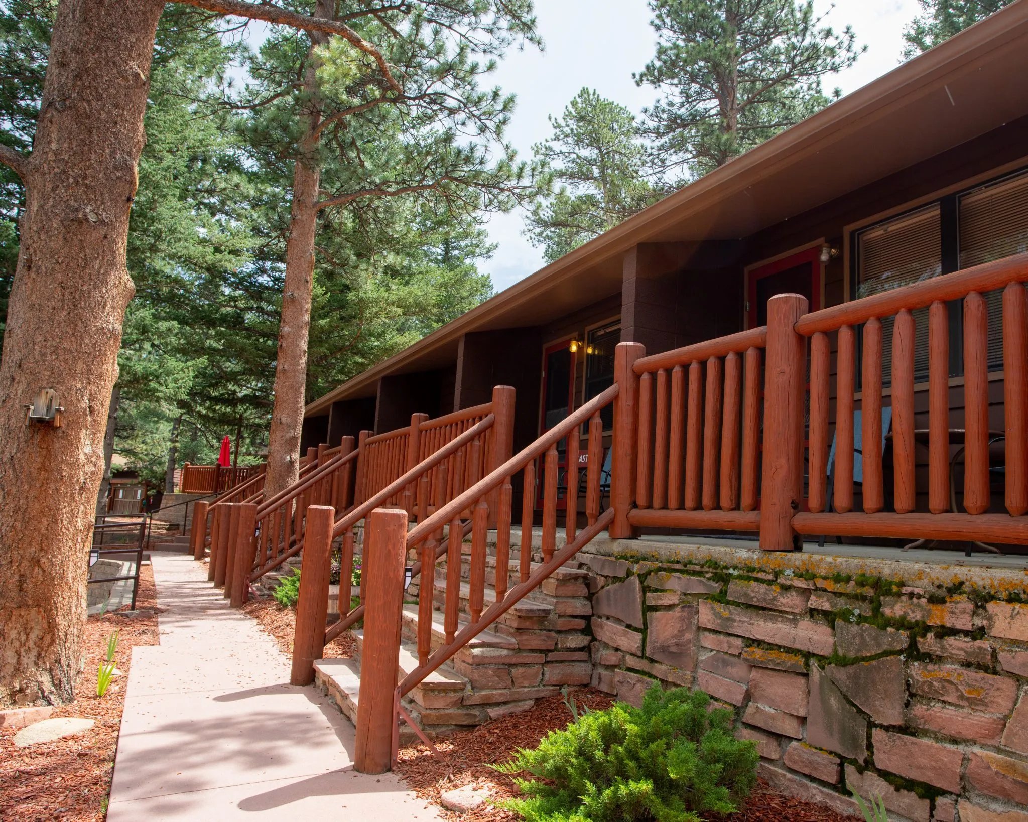 Long lodge building with multiple guest room entrances and log railings nestled in pine trees
