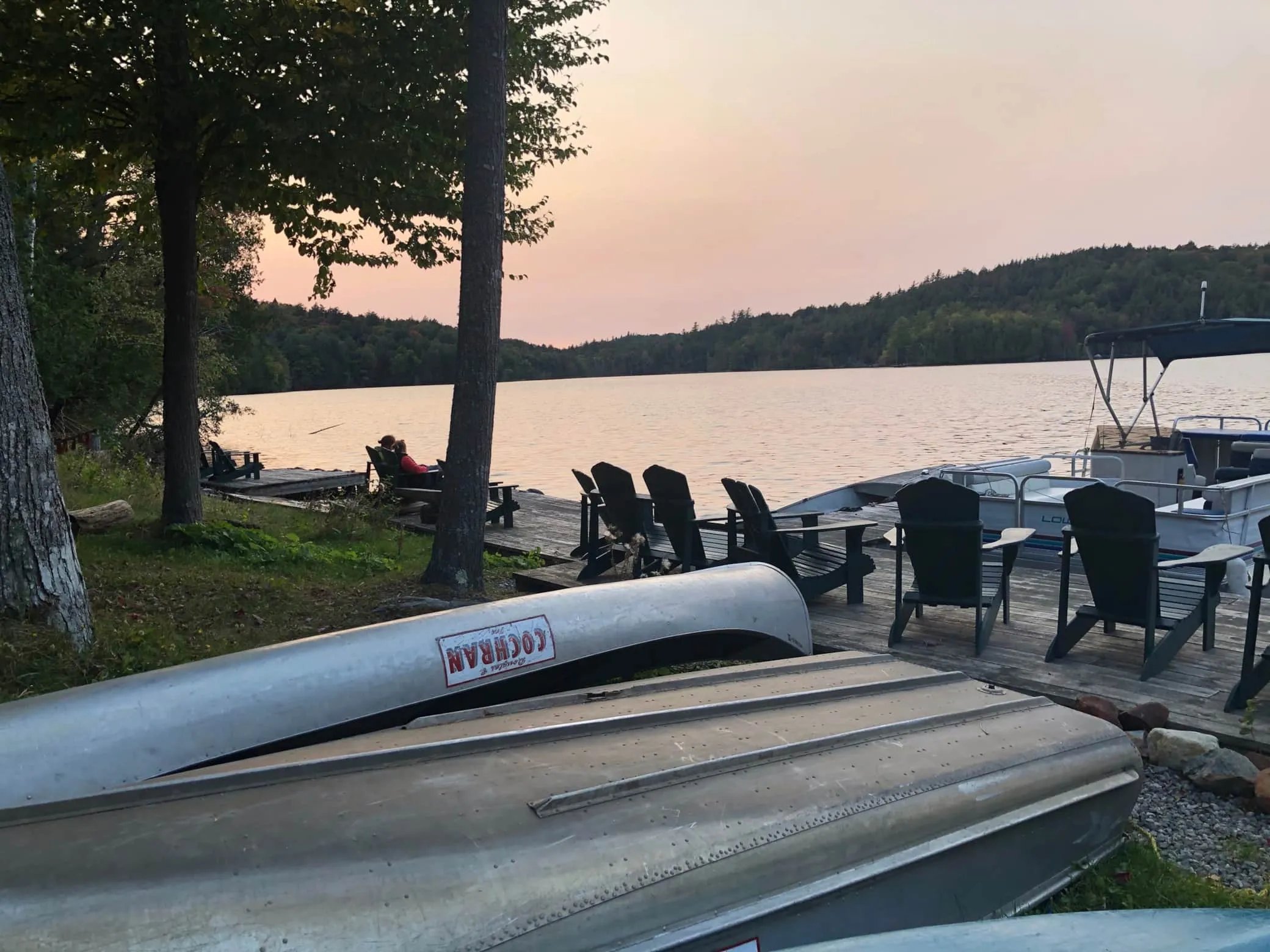 Canoes and Adirondack chairs on a lake shore at sunset.