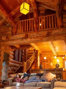 Log Cabin interior girl reading on sofa in living room with coffee table and 2nd story loft bedroom