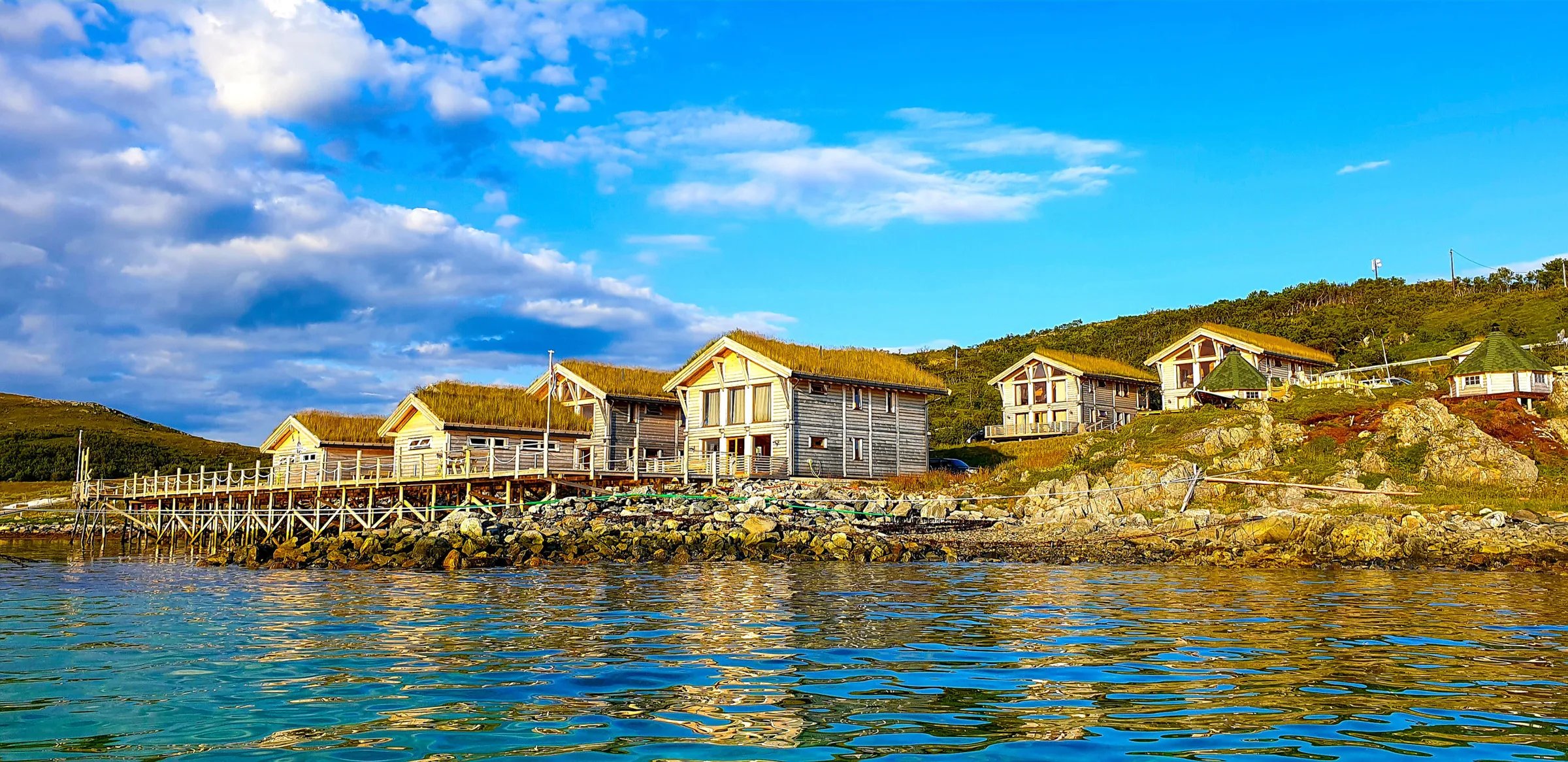 Lodge buildings with green roofs on a pier over clear blue water under a bright sky