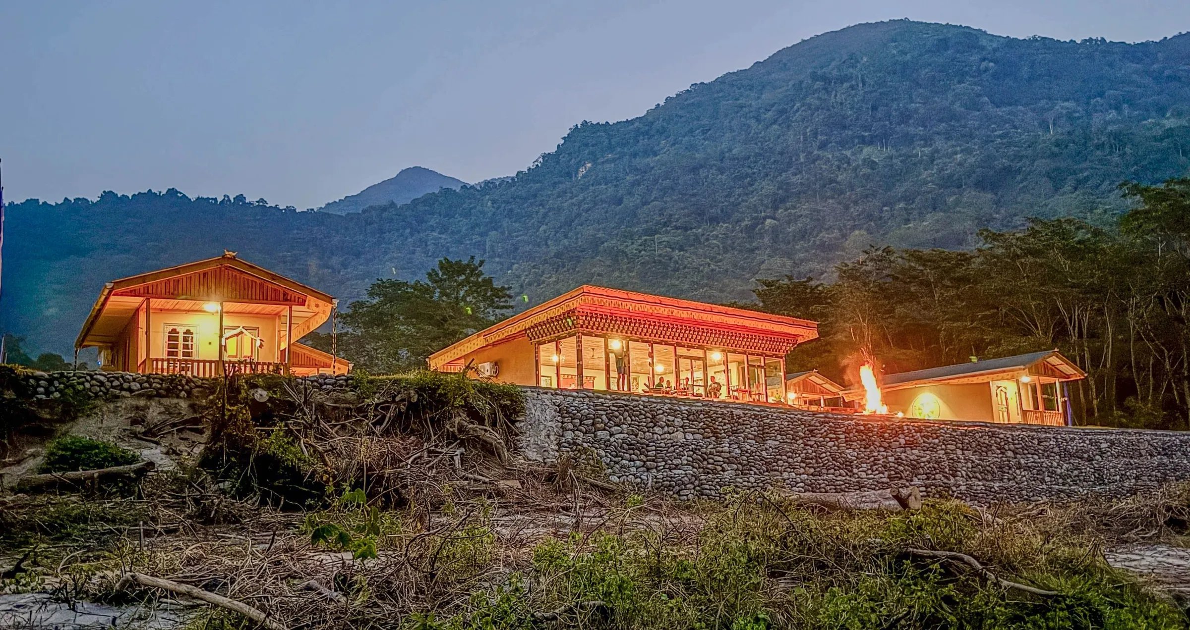 Manas River Lodge buildings illuminated at dusk with a campfire and mountain backdrop