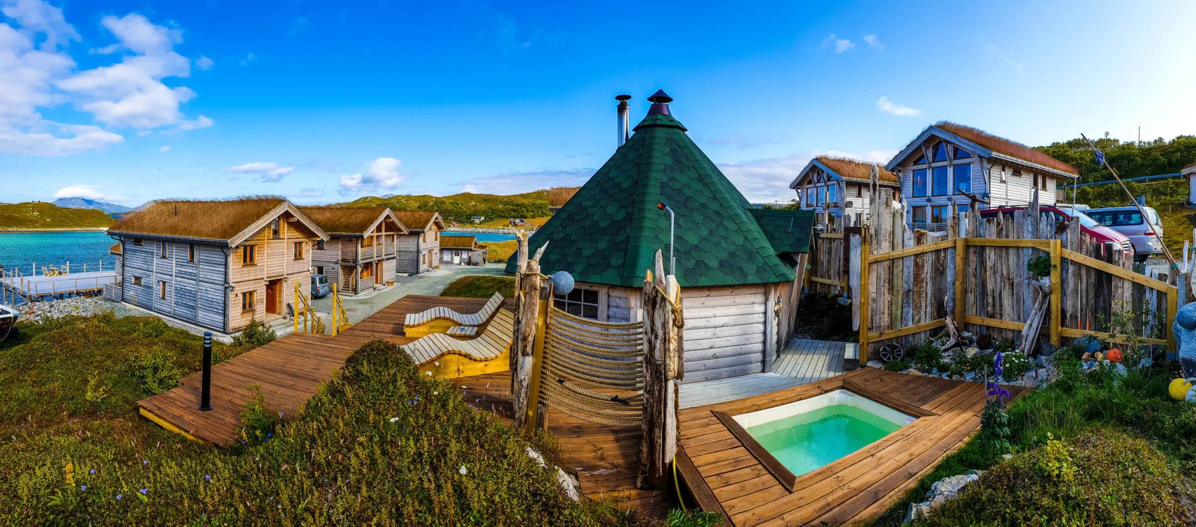 Aerial view of Mikkelvik Brygge lodge buildings on a pier over turquoise water, surrounded by green hills