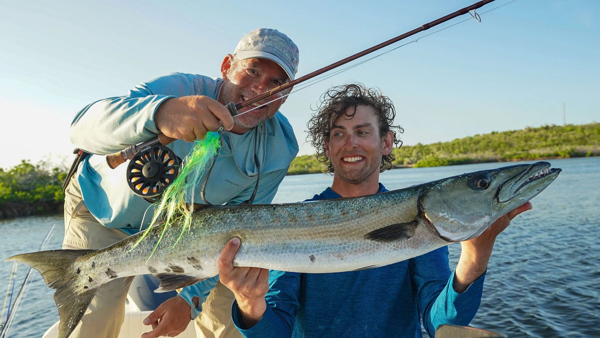 Two smiling anglers holding a large barracuda with a fly rod visible