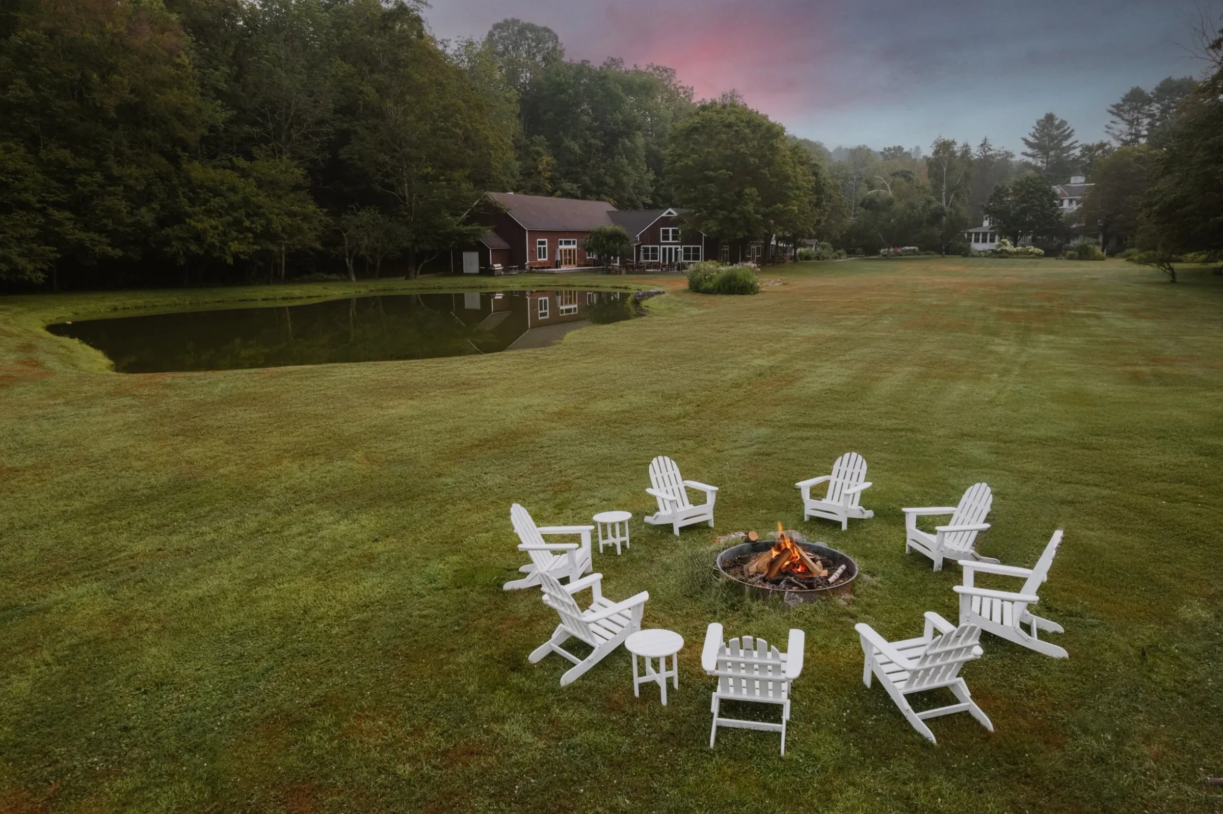 A circular arrangement of white Adirondack chairs around a small campfire on a grassy lawn with houses and trees in the background.