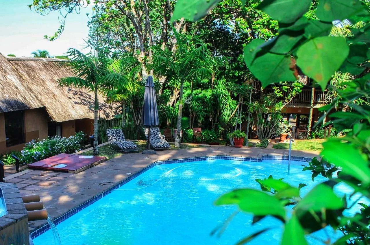 Thatched-roof lodge exterior and swimming pool illuminated at nightfall with palm trees