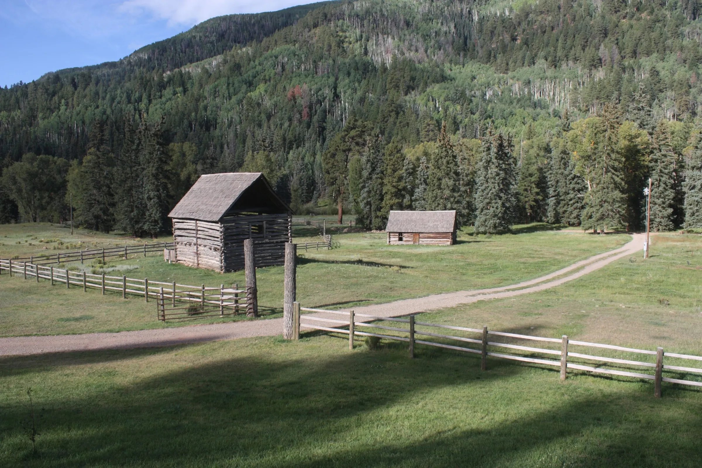 Two rustic wooden cabins or barns in a fenced green field with a dirt road, surrounded by forest
