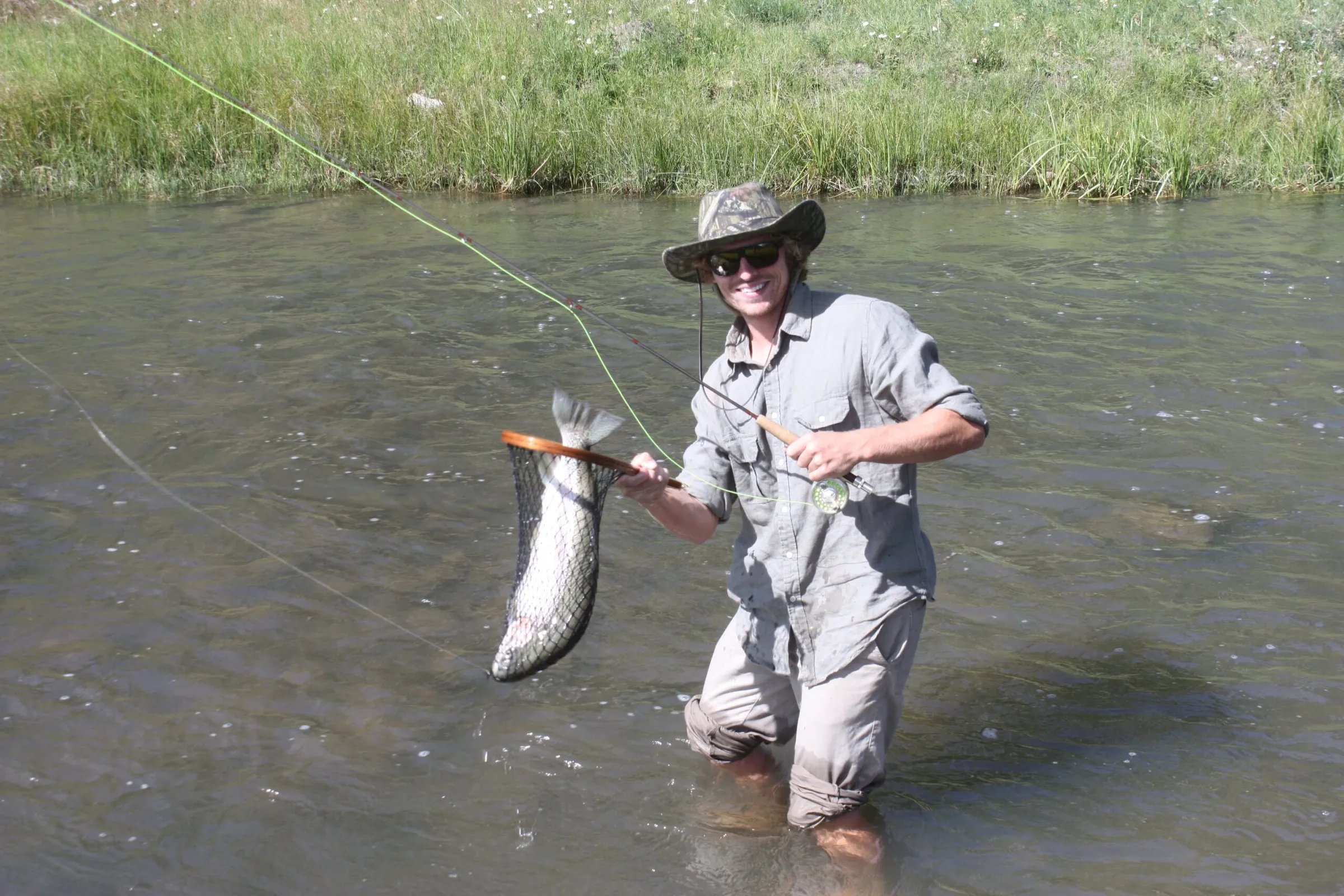 Angler in a hat holding a large fish in a net while standing in a shallow stream