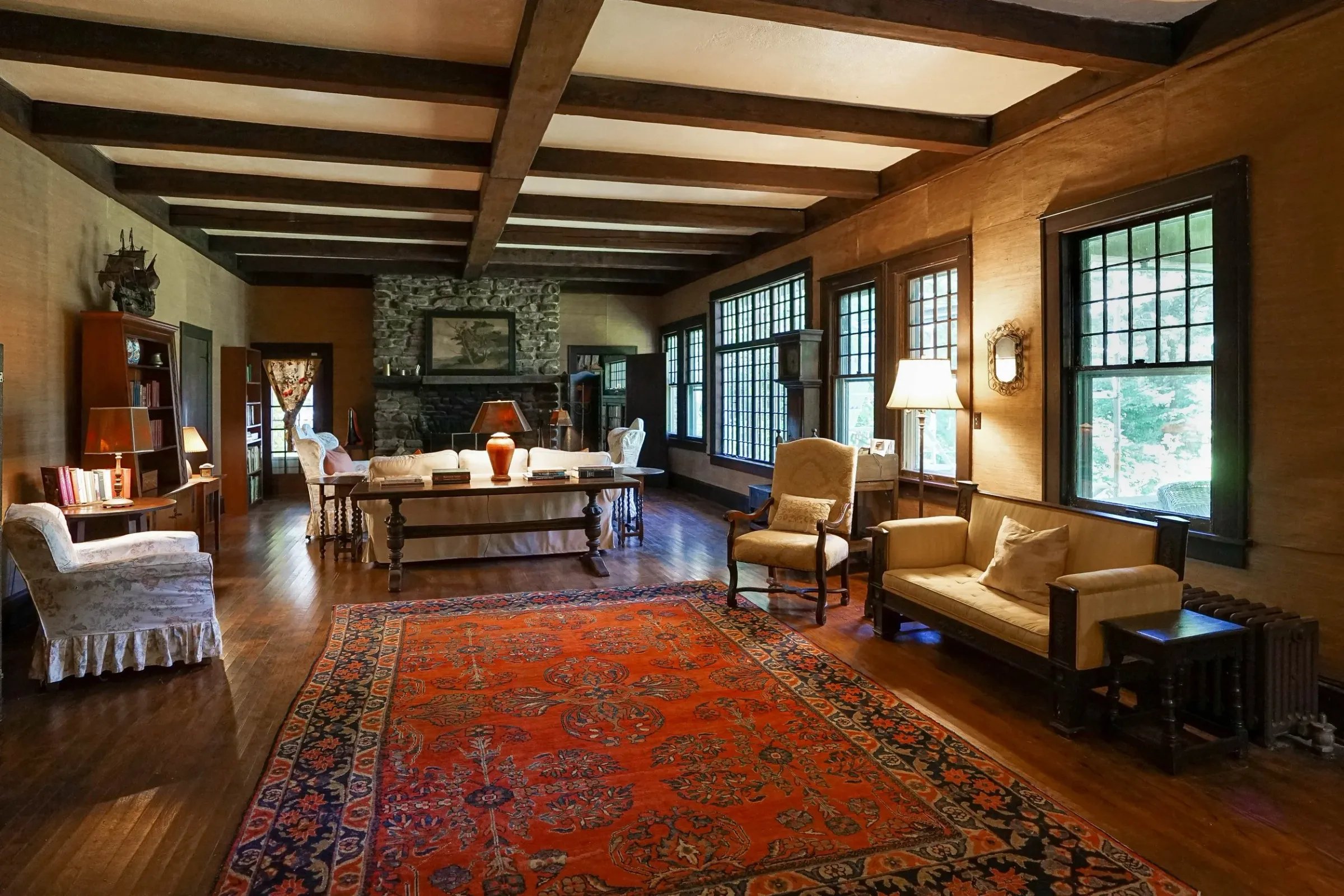 Living room with wooden floors, large windows, a stone fireplace, vintage furniture, and an orange patterned rug.