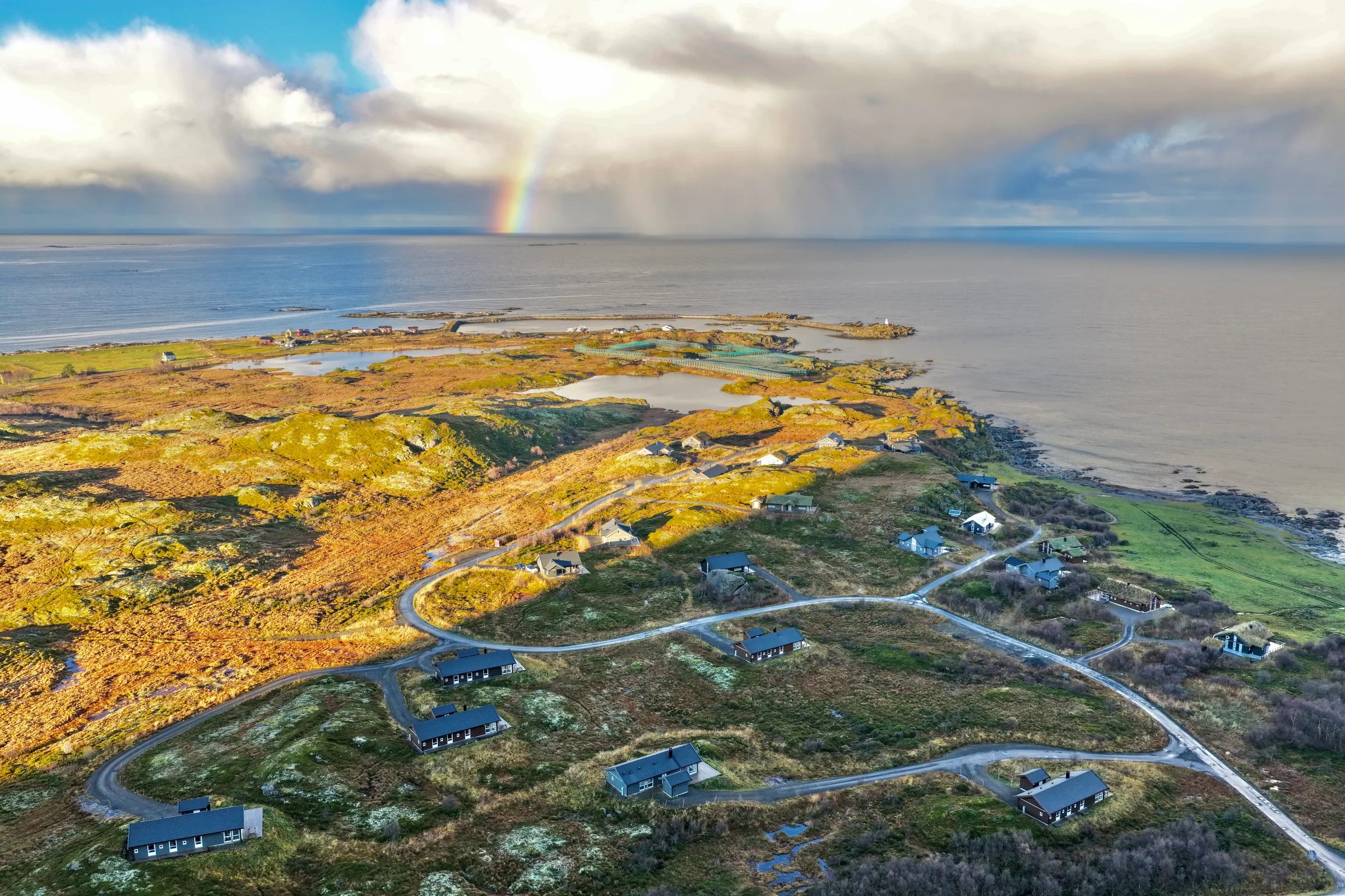 Aerial view of multiple lodges scattered across a green landscape with a rainbow over the ocean