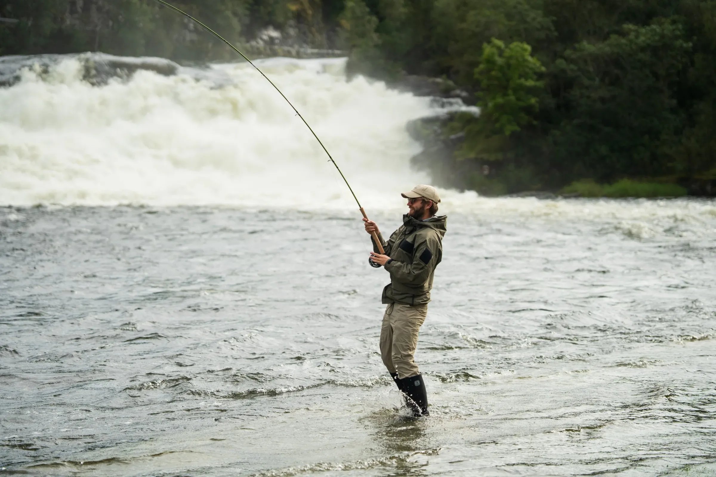 Angler wading in a river at sunset with forested mountains in the background.