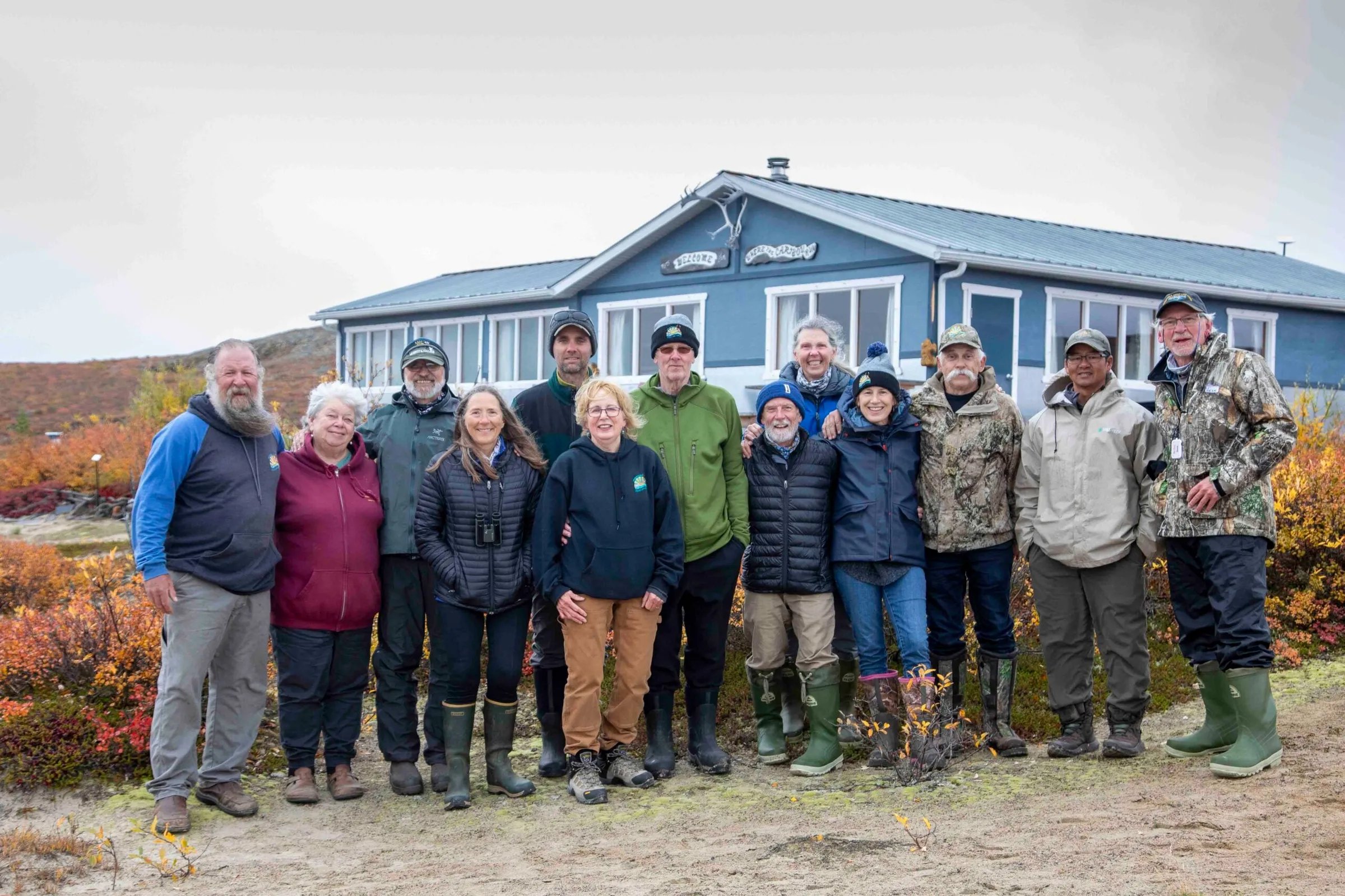 Group of staff and guests smiling in front of the blue lodge building on the tundra.