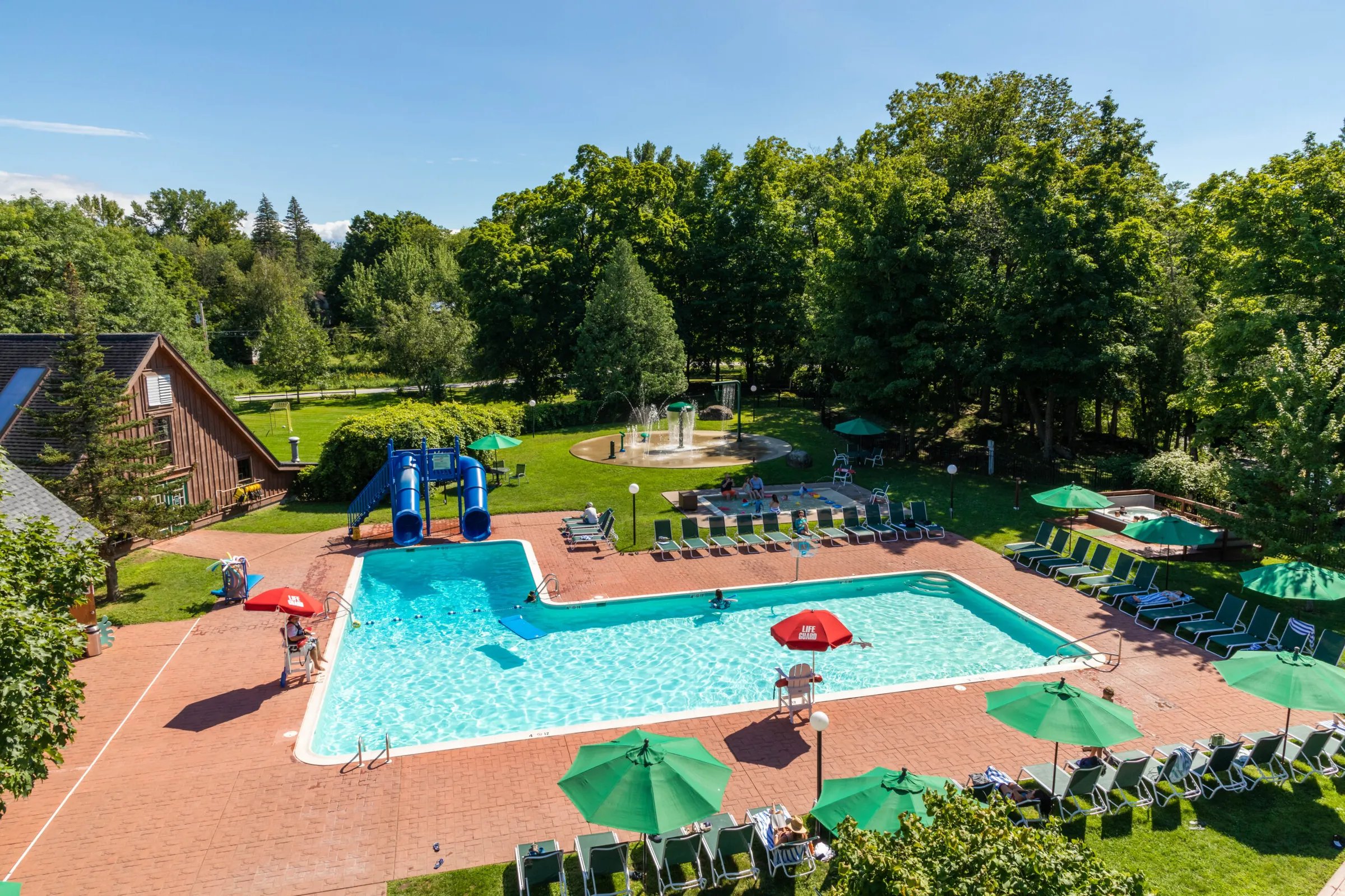 Aerial view of a large outdoor swimming pool complex and green grounds on a sunny day