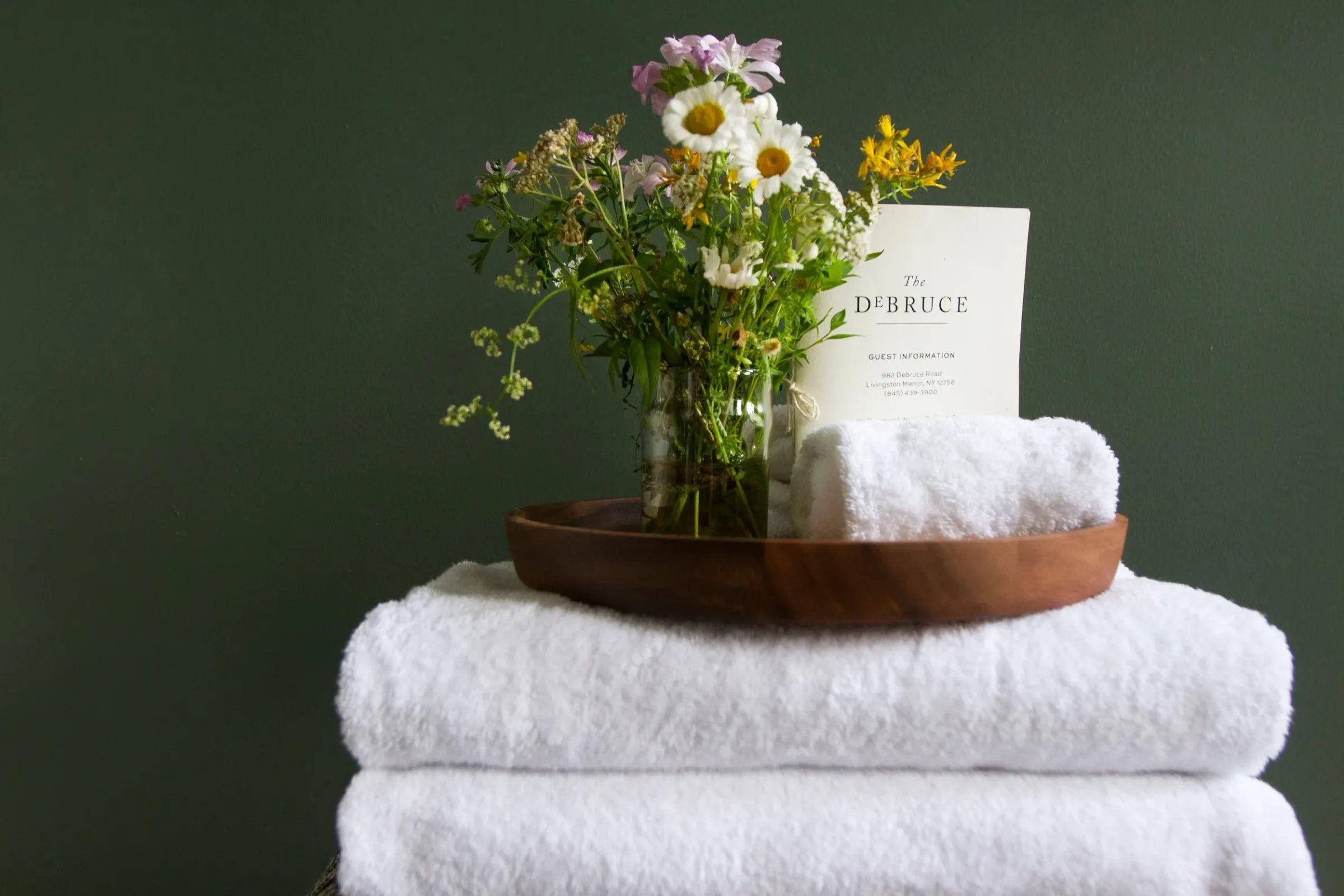 A wooden tray holding a rolled-up white towel and a glass vase with assorted fresh flowers, placed on top of two folded white towels, with a guest information card behind the arrangement, against a dark green wall.