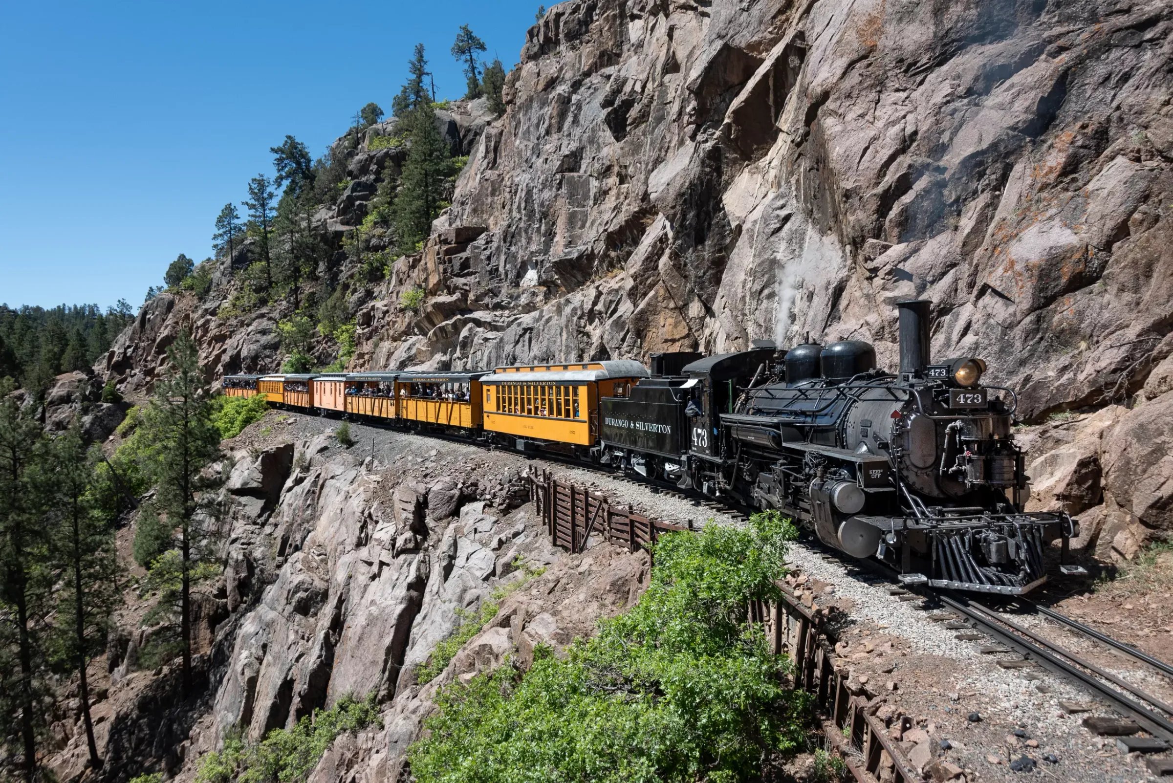 Durango & Silverton Narrow Gauge Train on a track cut into a rocky mountain face under a blue sky.