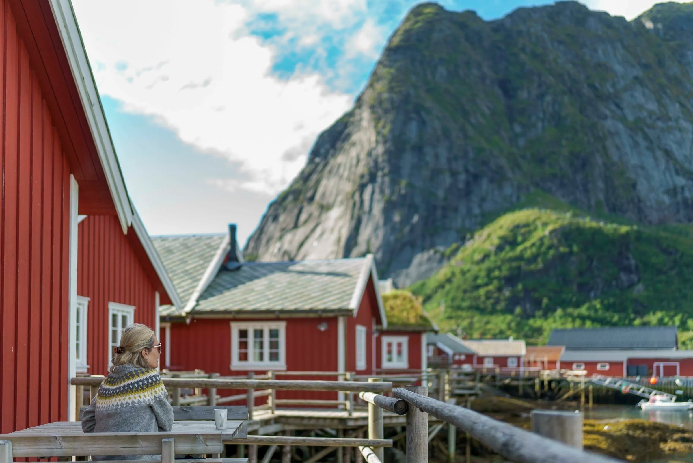 Person in a sweater sitting on a wooden deck of a red rorbu cabin, enjoying the mountain view