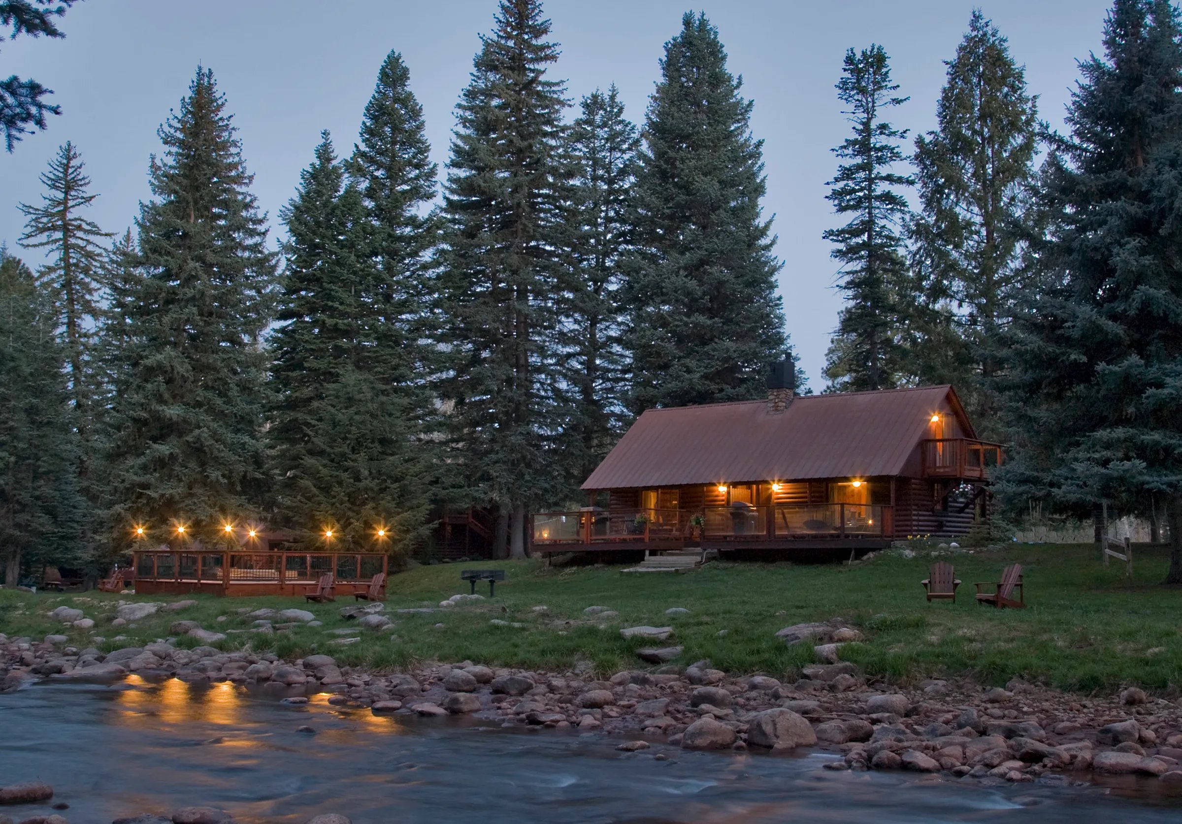 Log cabin by river at night with string lights reflecting on the water