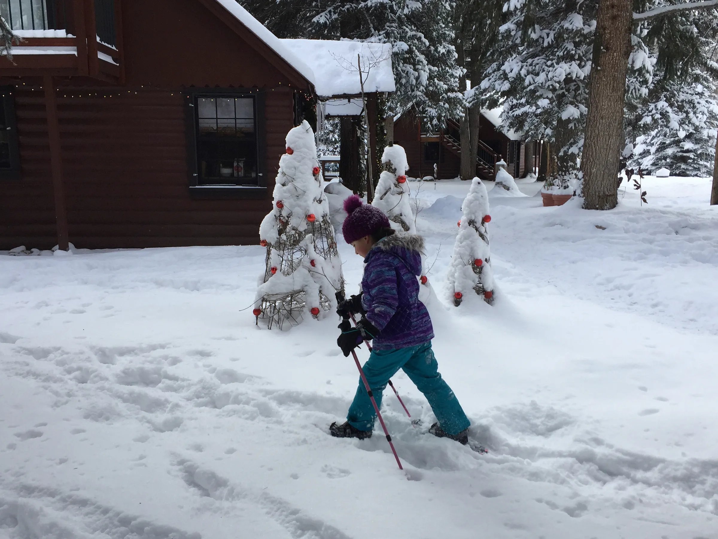 Child snowshoeing in front of a snow-covered cabin with decorated trees