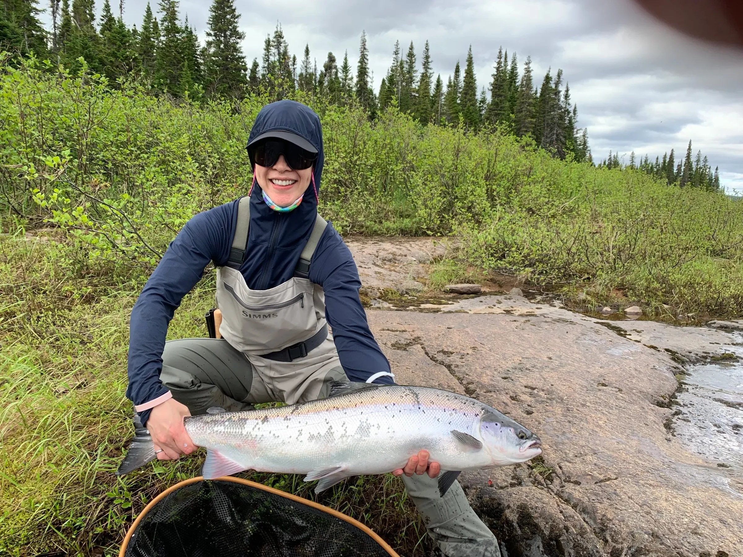 Angler smiling while holding a large Atlantic salmon on a rocky riverbank